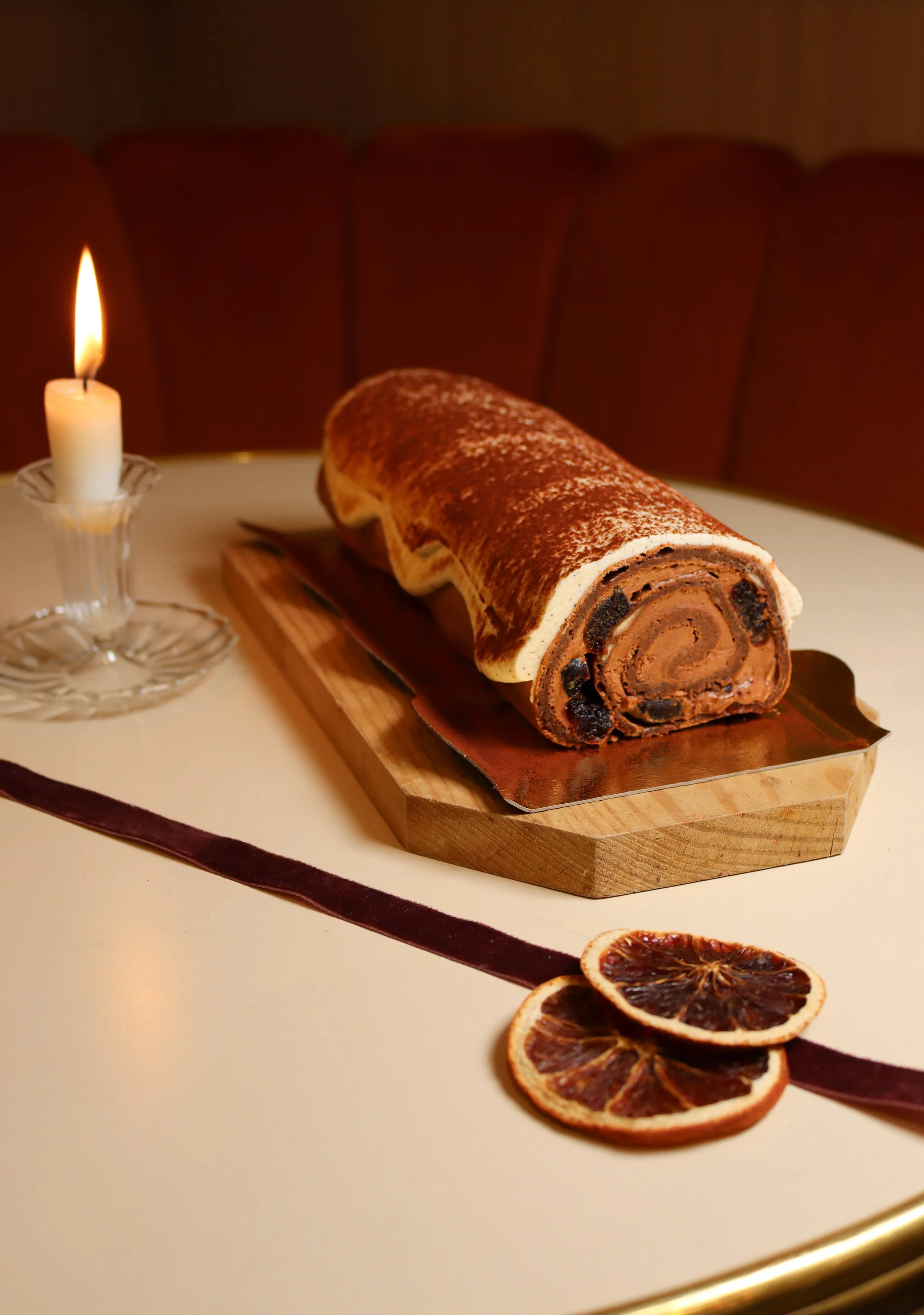 Gâteau roulé au chocolat avec raisins secs, disposé sur une planche en bois, avec des tranches d'orange séchée à côté, et une bougie allumée à gauche, sur un fond sombre.