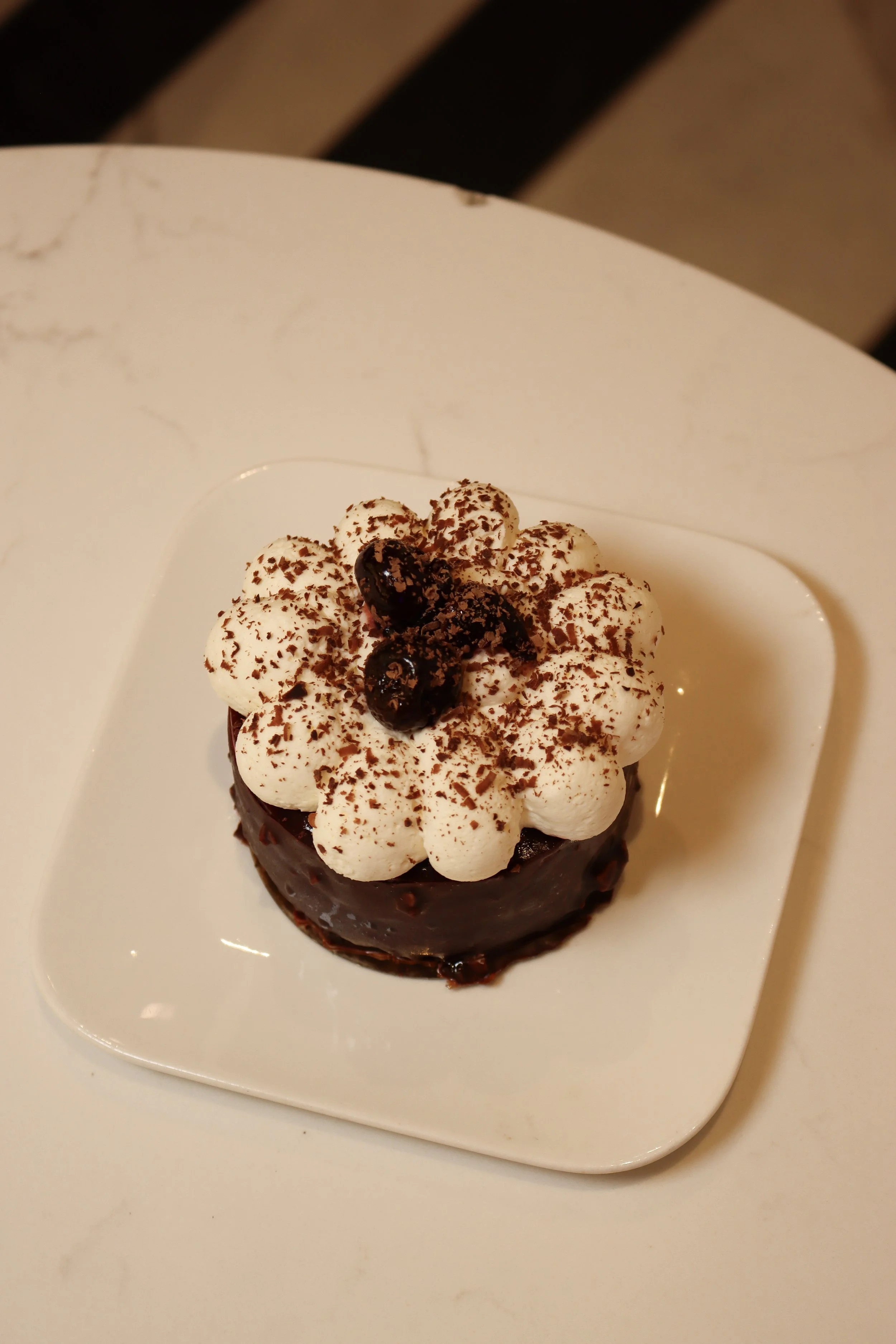 Gâteau au chocolat décoré de crème fouettée, cerises noires et chocolat râpé sur une assiette blanche.