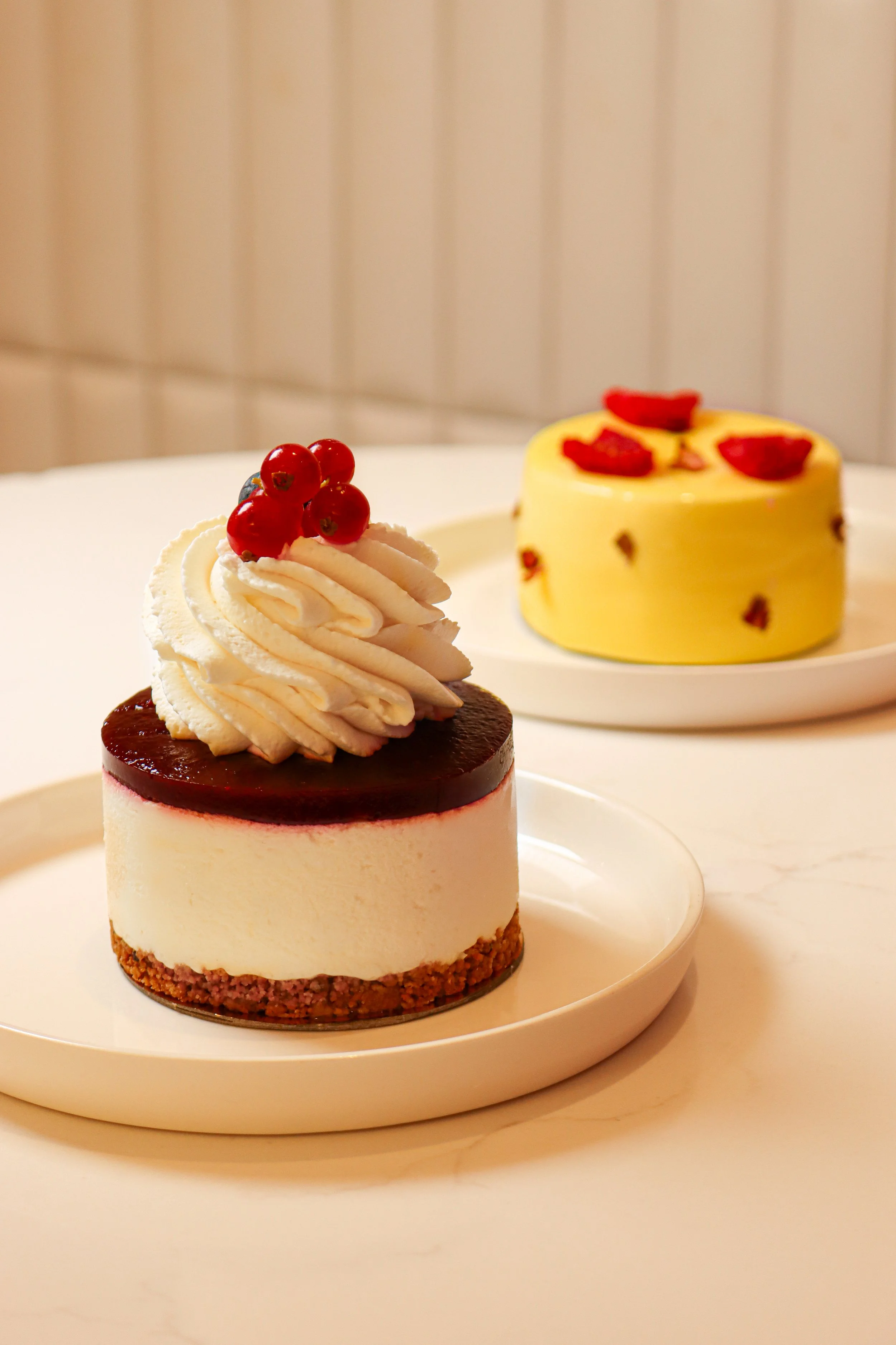 Un gâteau en forme de cylindre, avec une base à croquant, une couche de mousse blanche, une couche de gelée rouge, décoré d'une crème en spirale avec des groseilles rouges en haut, et un second gâteau en arrière-plan, également en forme de cylindre avec des groseilles et des morceaux rouges sur le dessus.