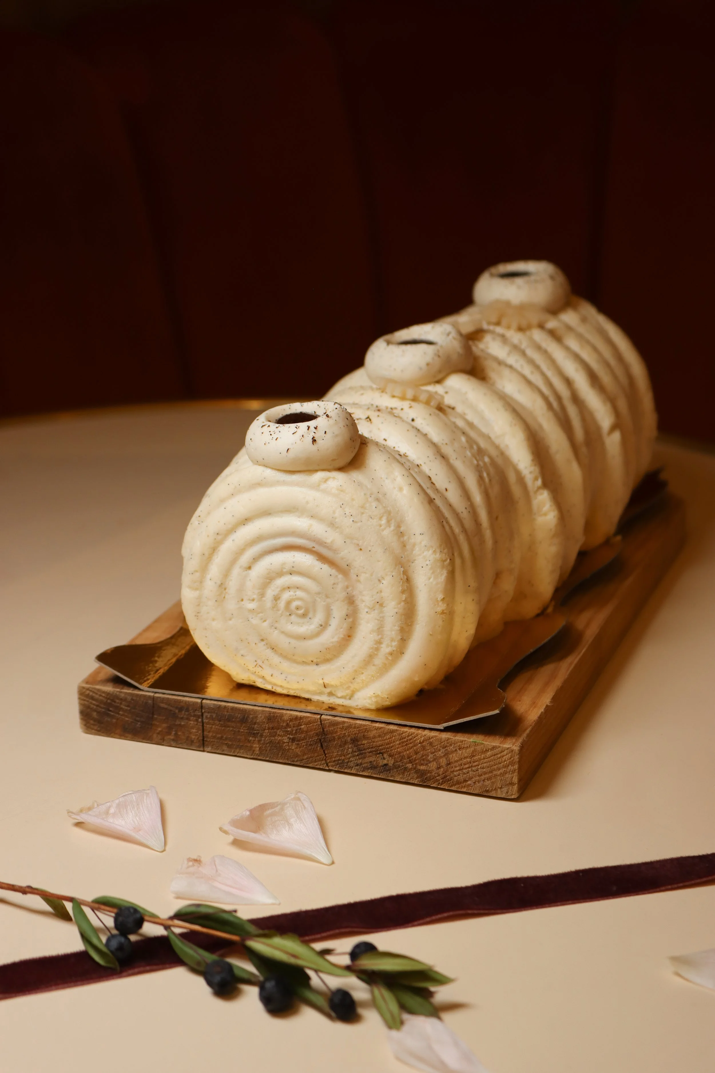 Gâteau roulé à la vanille avec décoration de fondant blanc et petits disques blancs en haut, placé sur une planche en bois, avec des pétales de fleurs et des branches avec des baies en premier plan.