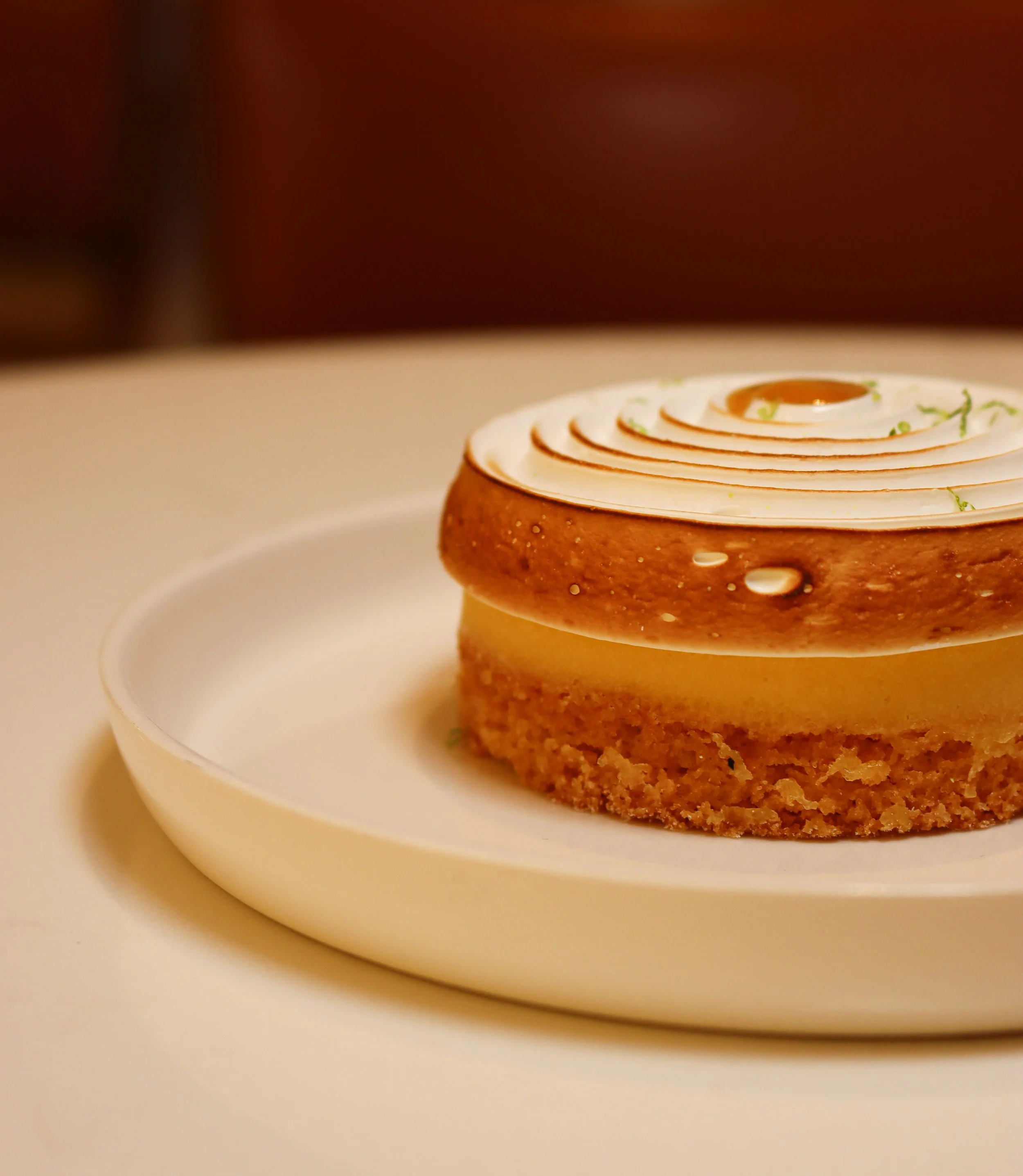 Gâteau rond avec glaçage blanc, décoré de spirales de crème et d'une fine couche de zestes de citron vert, présenté sur une assiette blanche.