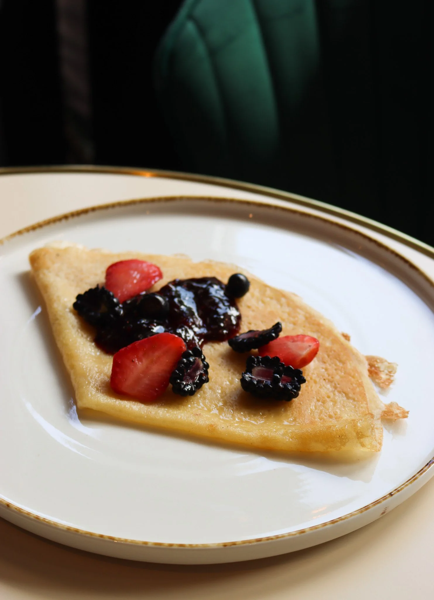 Une tranche de gâteau ou de crêpe garnie de fruits rouges, comme des fraises, des mûres et des cassis, servie sur une assiette blanche ornée d'une fine ligne dorée.