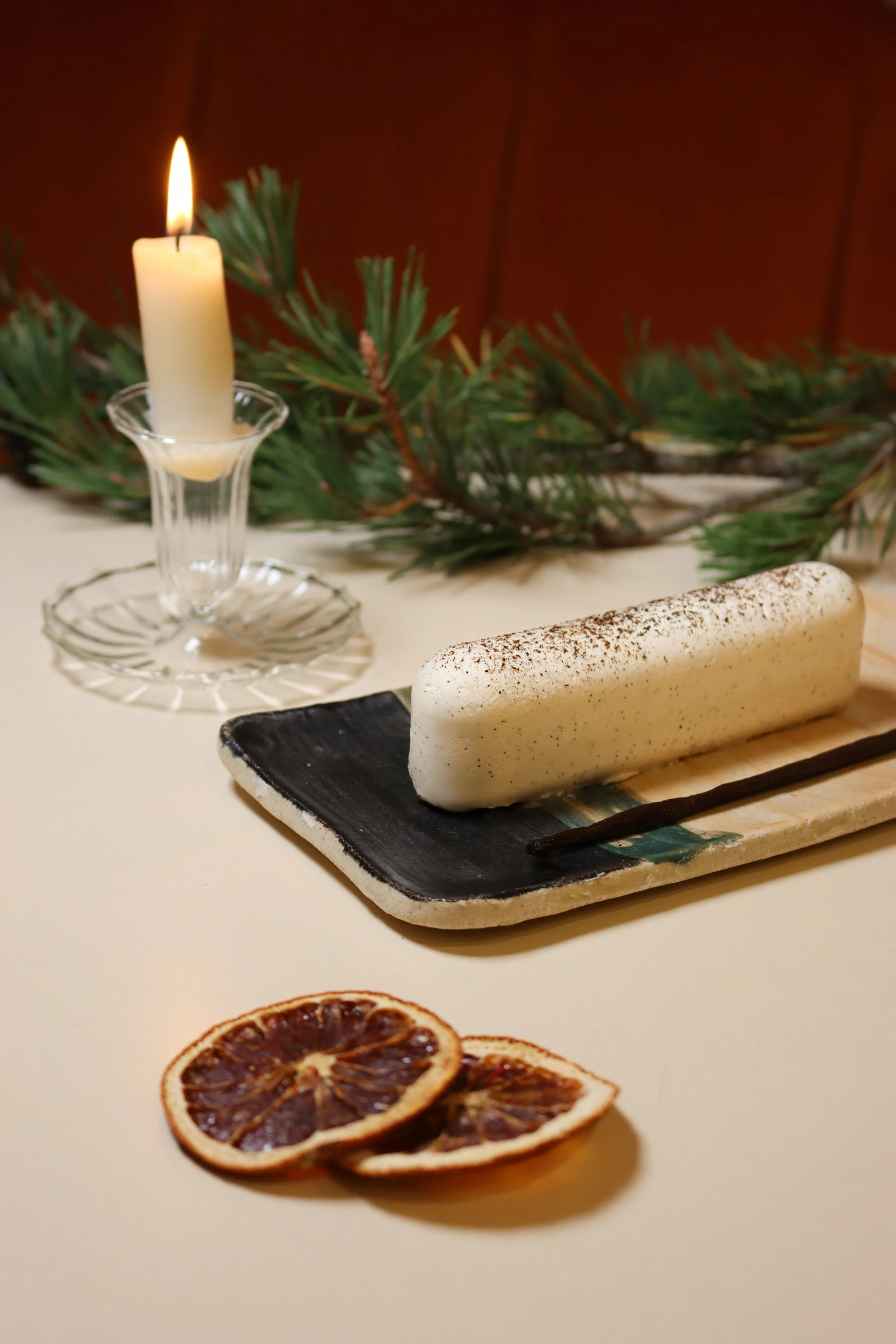 Une bougie blanche allumée dans un chandelier en verre, entourée de branches de sapin, sur une table avec un gâteau blanc, et en arrière-plan on voit des tranches de citron confit.