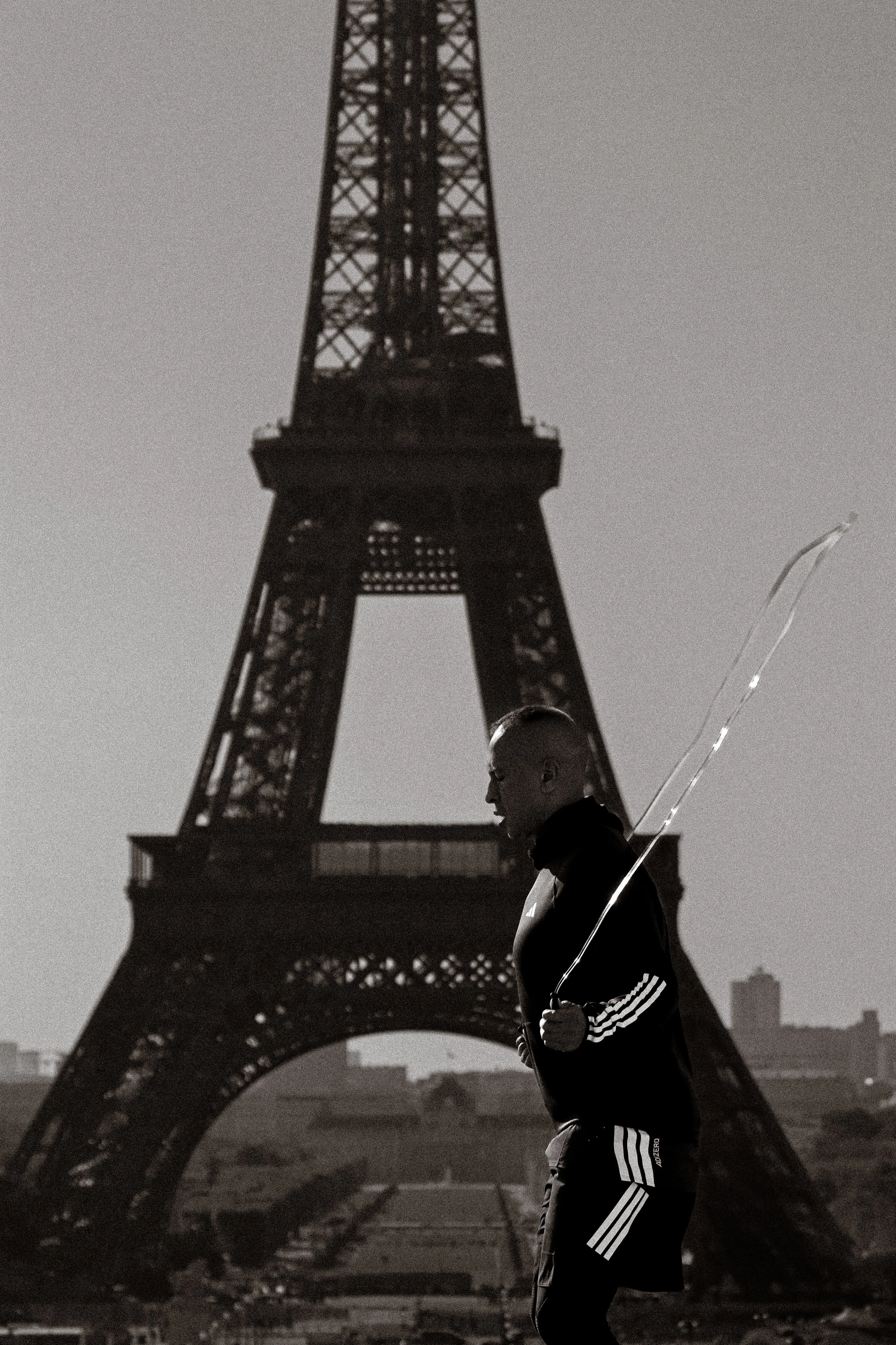 Photographie de sport: Une personne en jogging noir avec des rayures blanches sur les côtés fait de la corde à sauter devant la tour Eiffel à Paris, en plein air, lors d'une journée nuageuse. photographie en noir et blanc.
