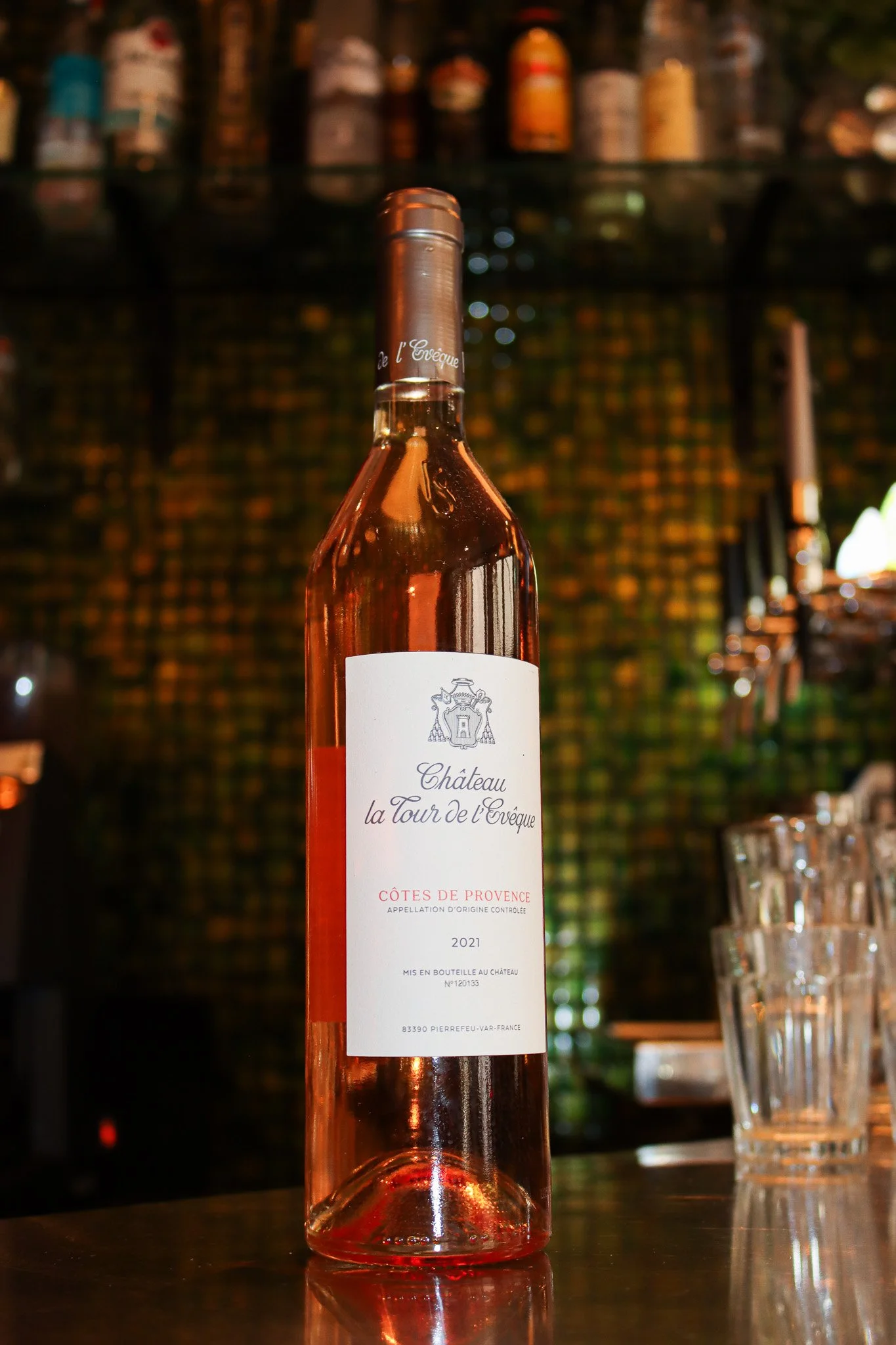 Bouteille de vin rosé Château la Tour de l'Evêque, 2021, dans un bar ou un restaurant, avec étagères de bouteilles en arrière-plan et un verre d'eau à côté.