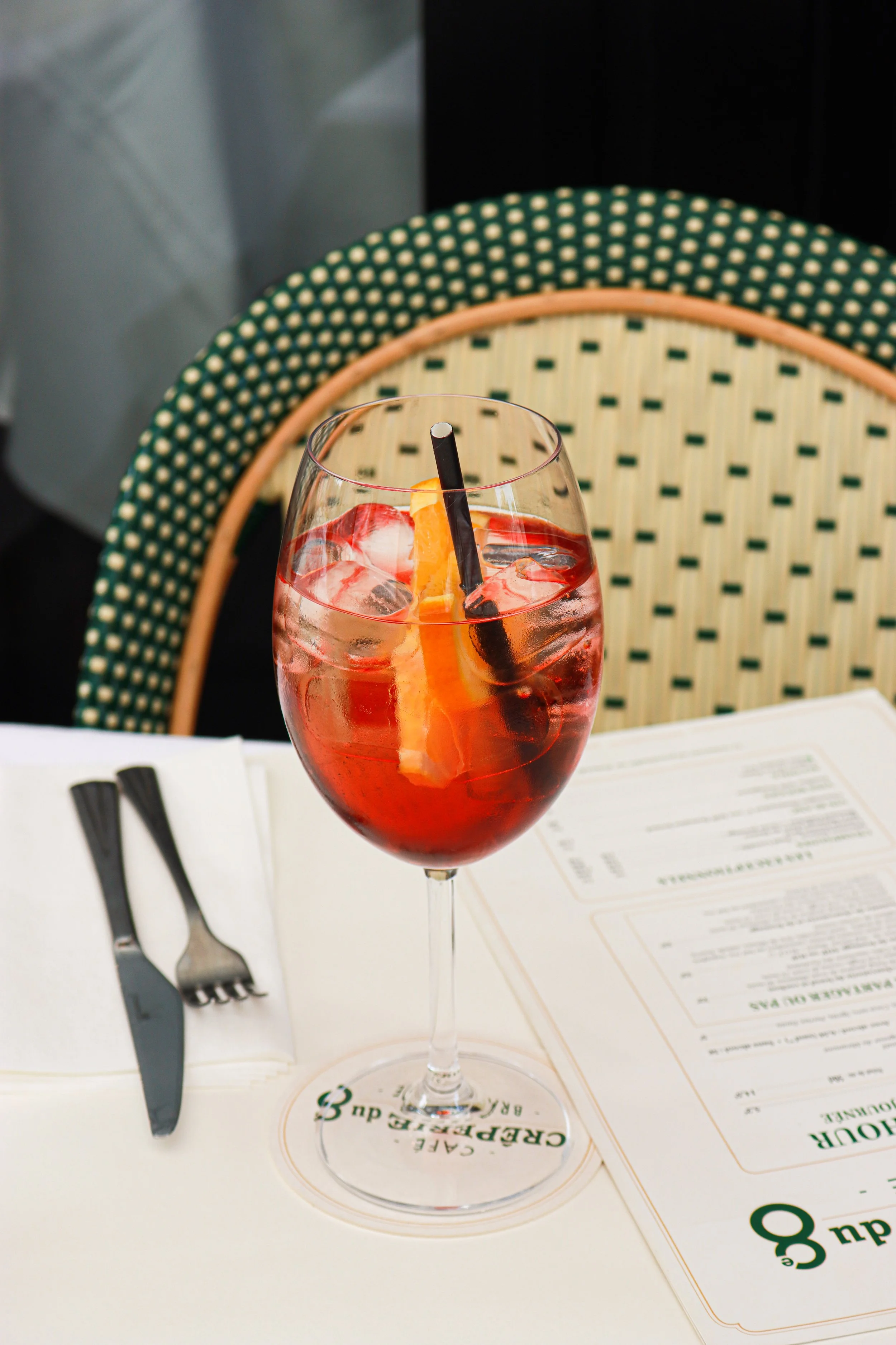Verre à vin rouge avec glaçons, une tranche d'orange et une paille noire sur une table avec un menu et deux fourchettes.