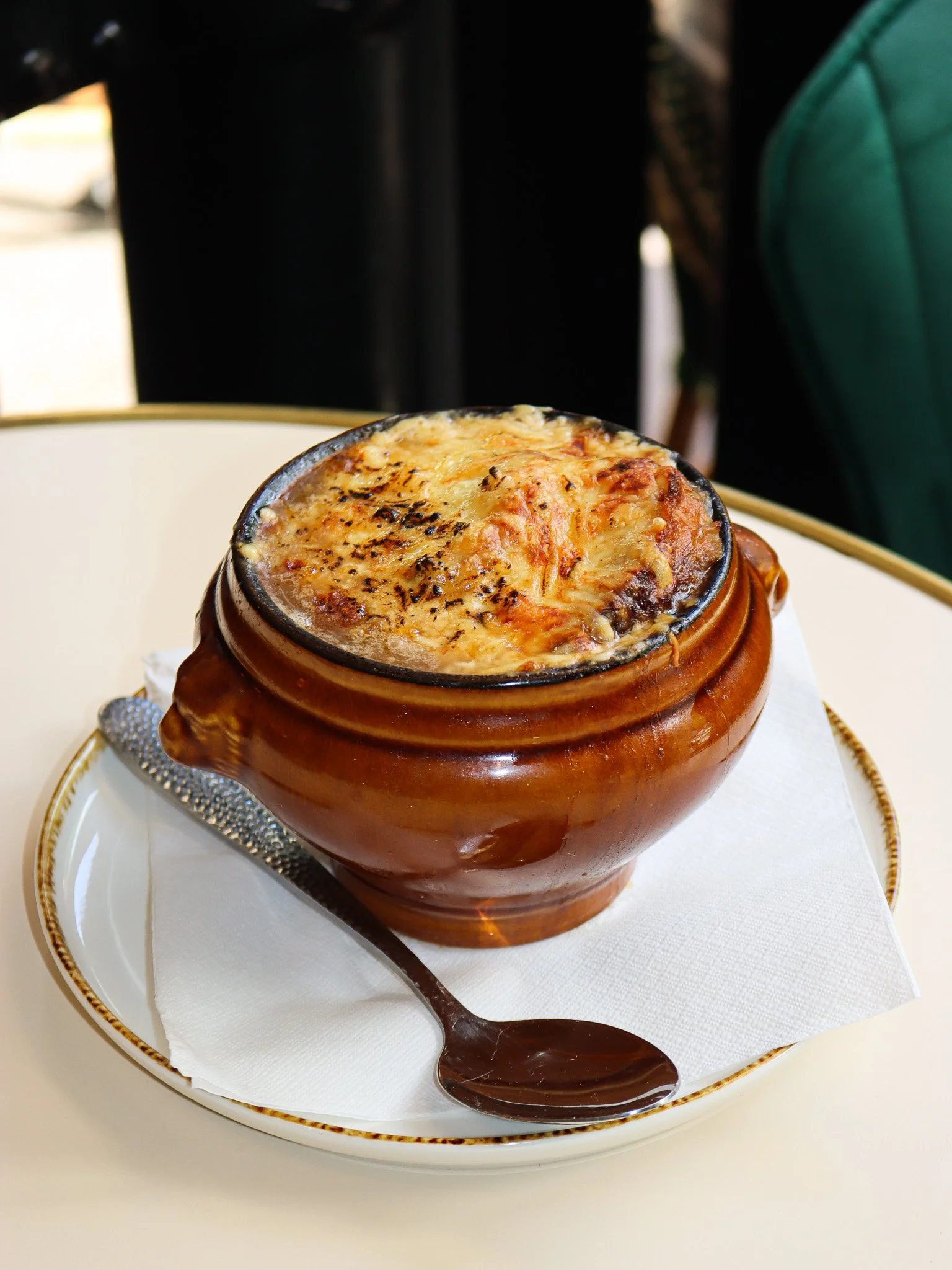 Cocotte en terre cuite avec un gratin de fromage, servi sur une assiette avec une cuillère en métal, sur une nappe blanche.