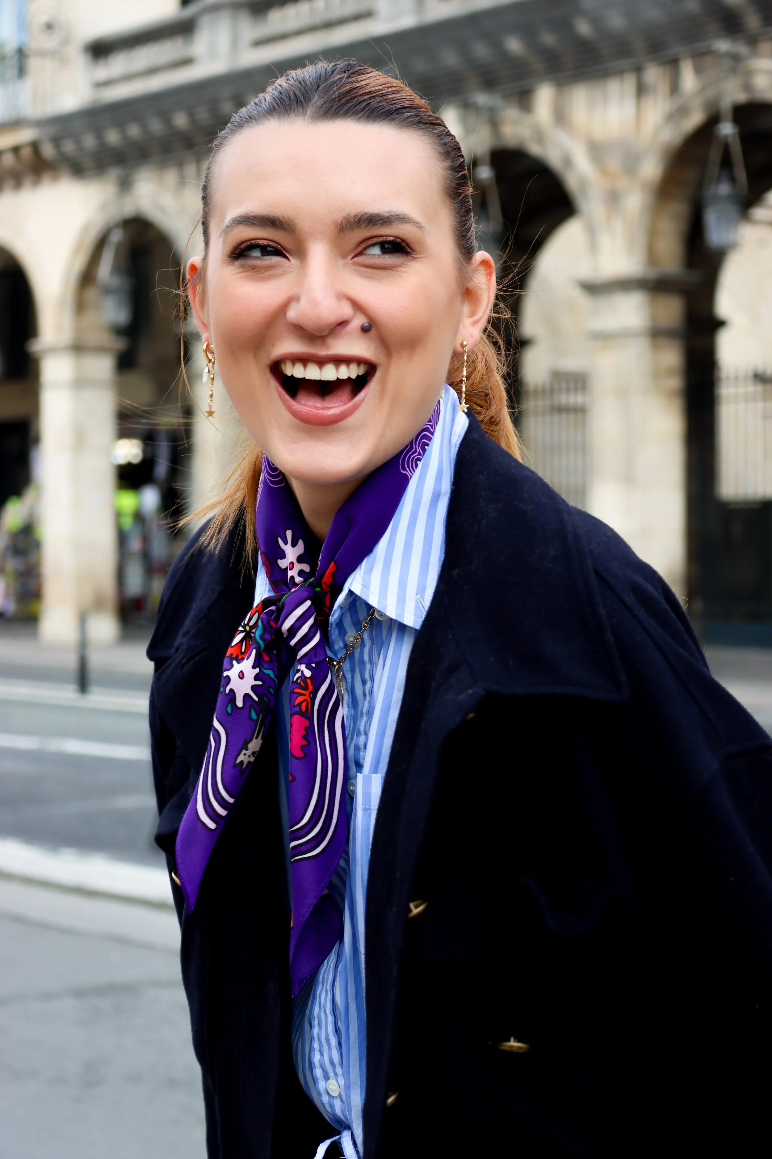 Une femme souriante avec une langue sortie, portant des boucles d'oreilles pendantes, un foulard violet avec des motifs, une chemise à rayures bleues et blanches, et un manteau noir, devant un bâtiment avec des arches en pierre.