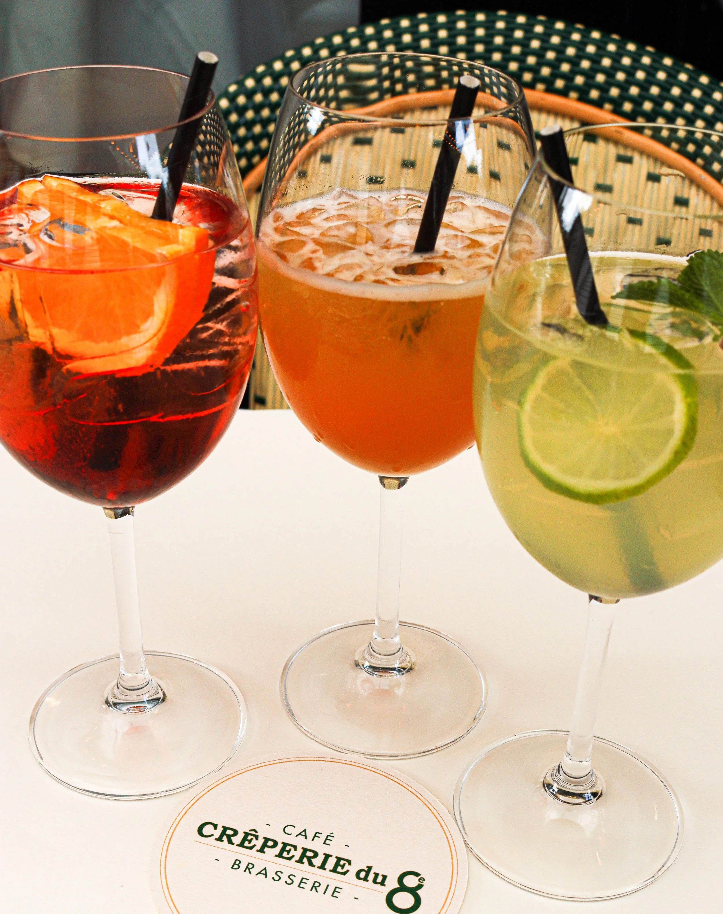 Trois verres de boisson colorée avec pailles, garnis de fruits comme citrons, oranges ou menthe, sur une table avec un menu ou une carte de café, crêperie, brasserie.