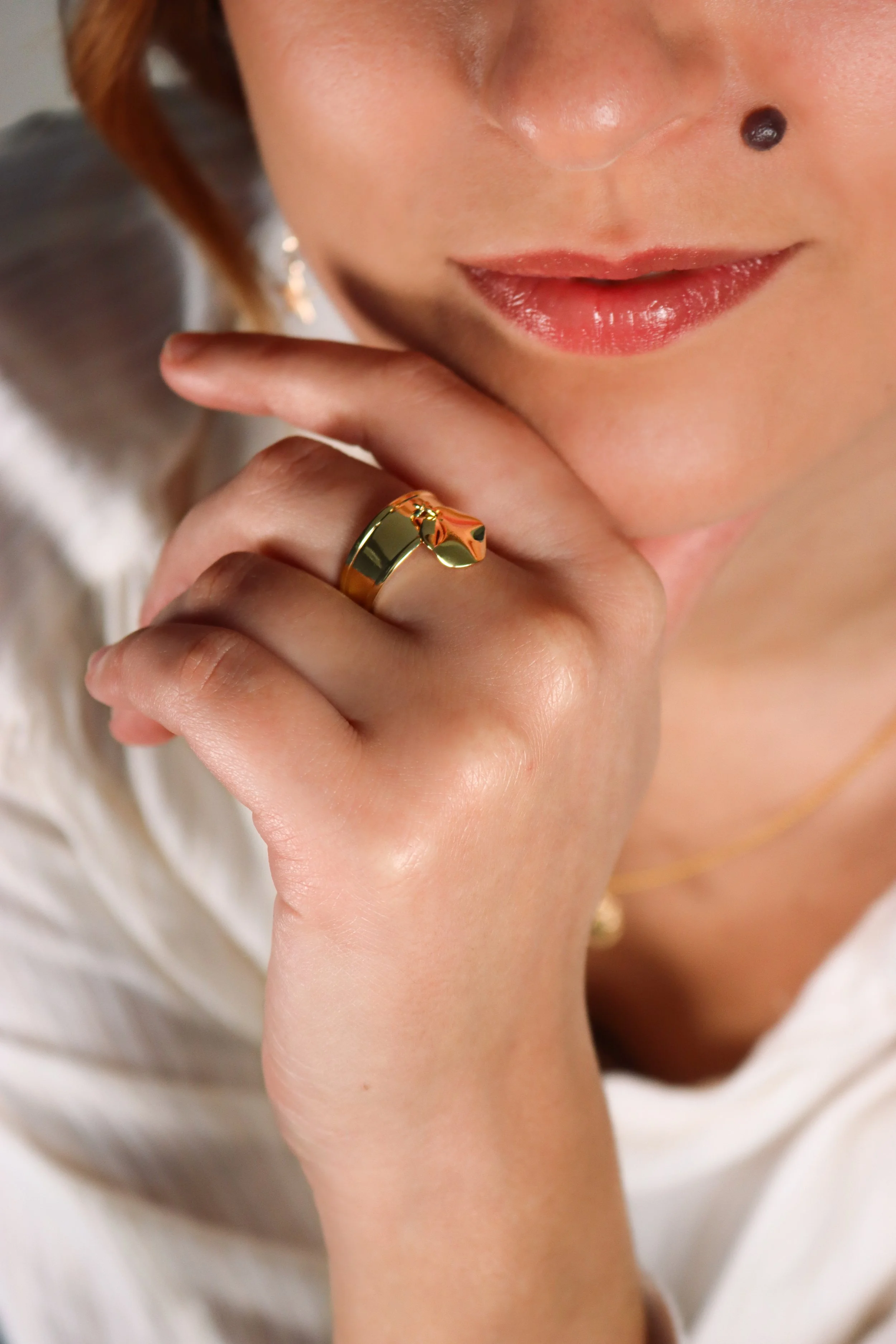 Gros plan du visage d'une femme avec un maquillage léger, portant un bague en or sur le doigt, avec une main près du visage, lèvres maquillées, et un piercing au nez.