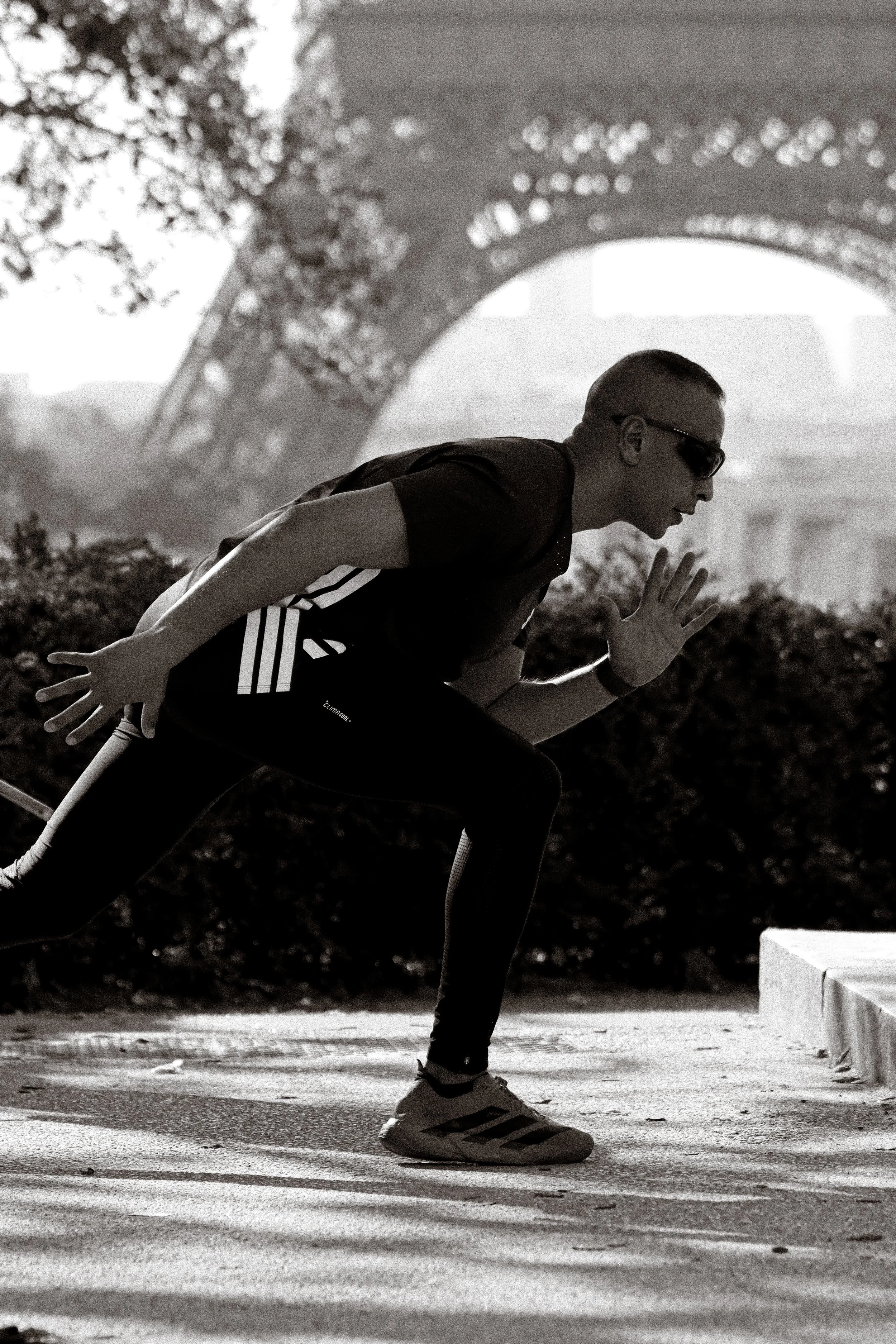 Photographie de sport: Un homme en position de sprint près à monter les escalier du Trocadéro à Paris, portant des lunettes de soleil, un t-shirt noir et des chaussures de sport, avec la Tour Eiffel en arrière-plan, en noir et blanc.