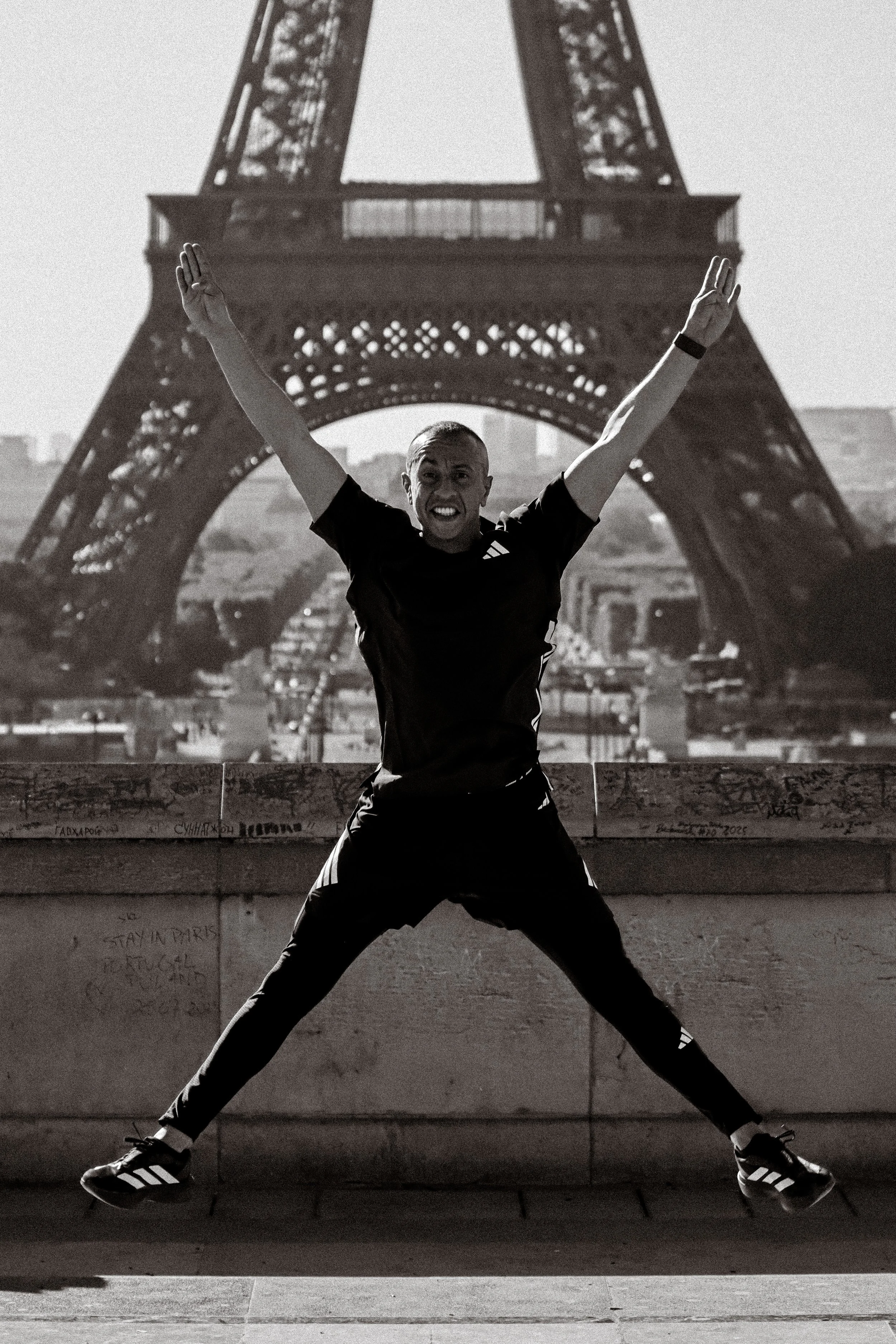 Une personne en position large, avec les bras levés, sur un mur, avec la Tour Eiffel en fond, en Paris, photographiée en noir et blanc.