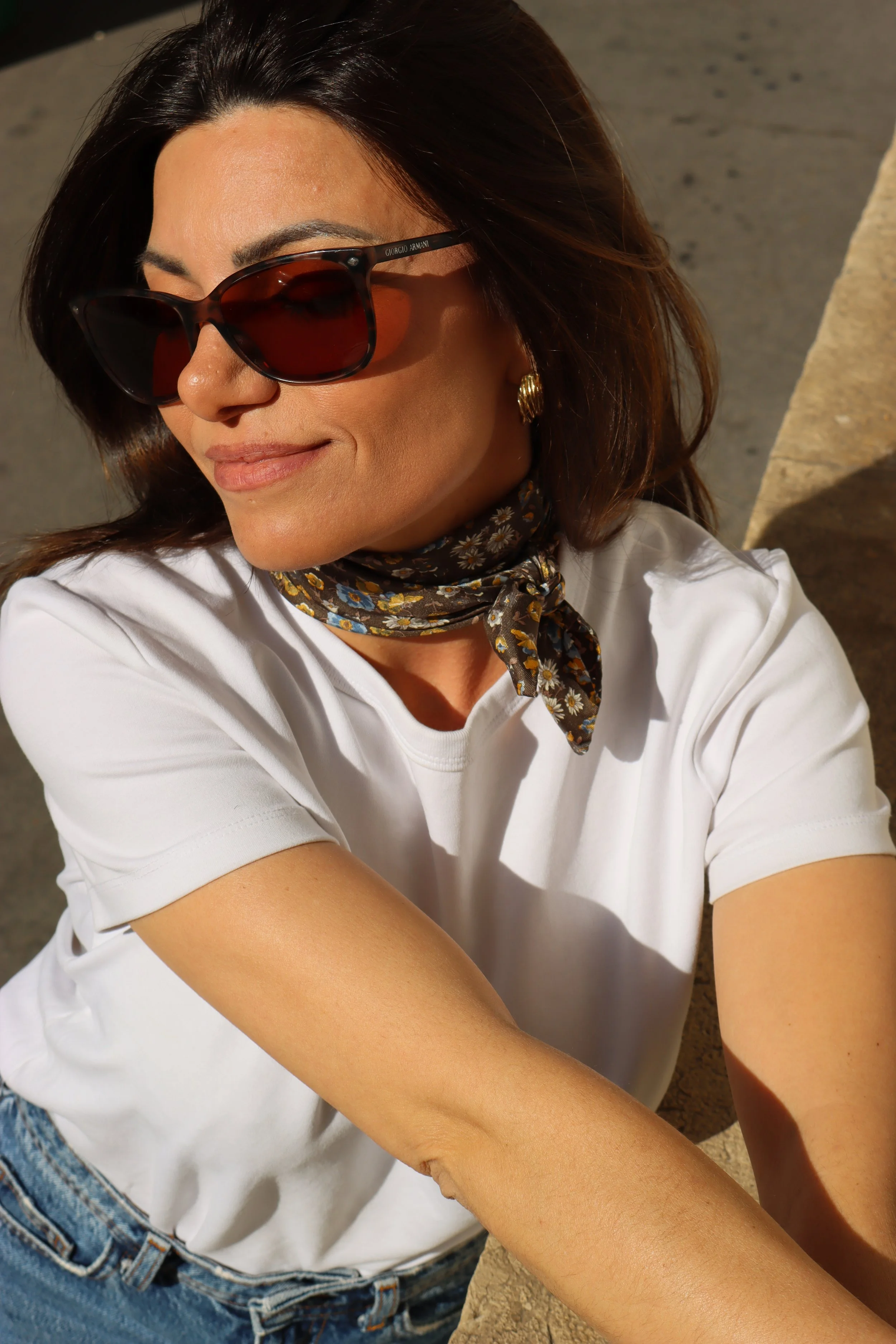 Une femme portant des lunettes de soleil, un foulard à motifs floraux autour du cou, une chemise blanche, et un jean, sourit devant un fond en pierre, avec la lumière du soleil qui illumine son visage.