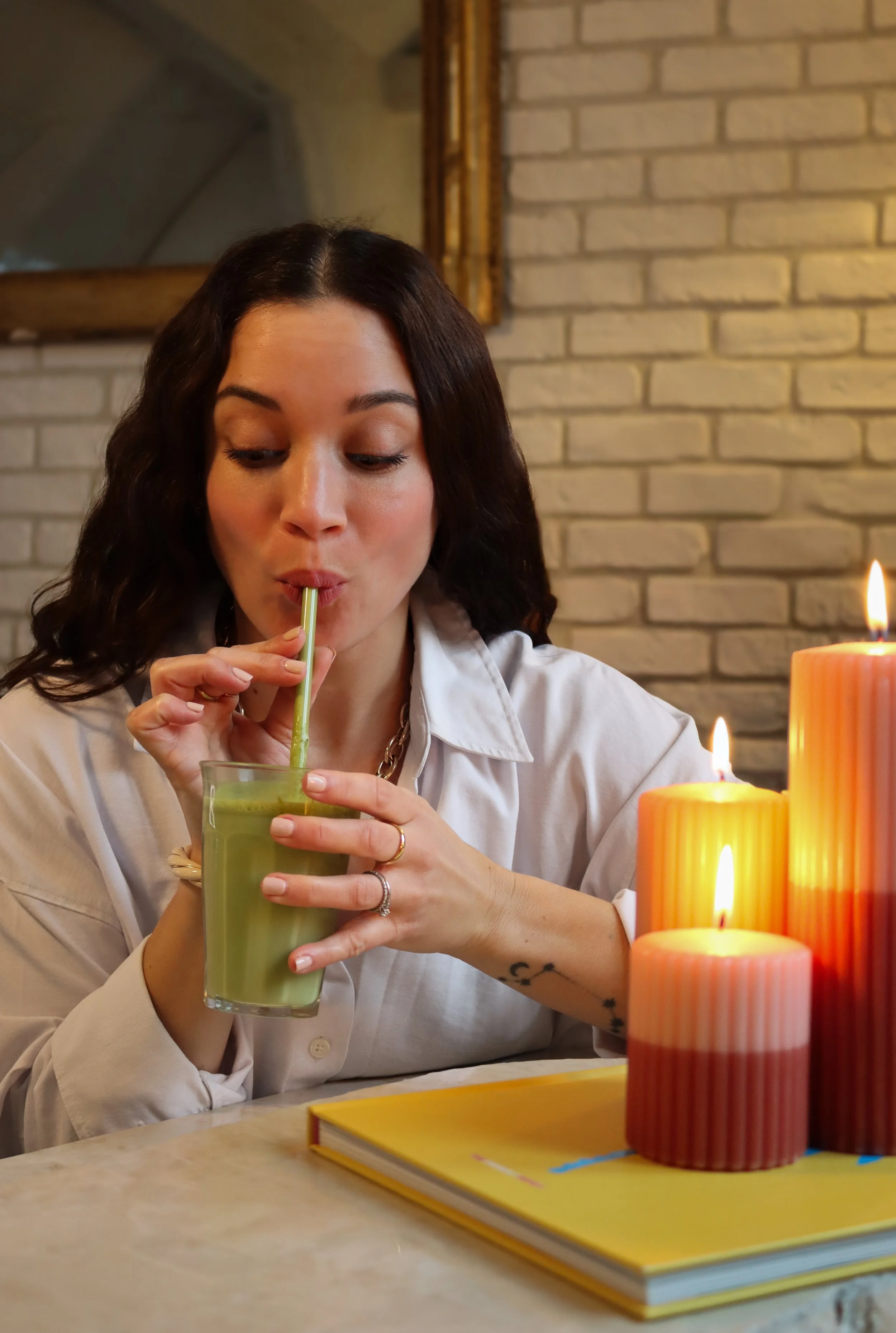 Une femme aux cheveux bouclés et à la peau claire boit un smoothie vert à l'aide d'une paille, assise devant une cheminée en brique et des bougies allumées.