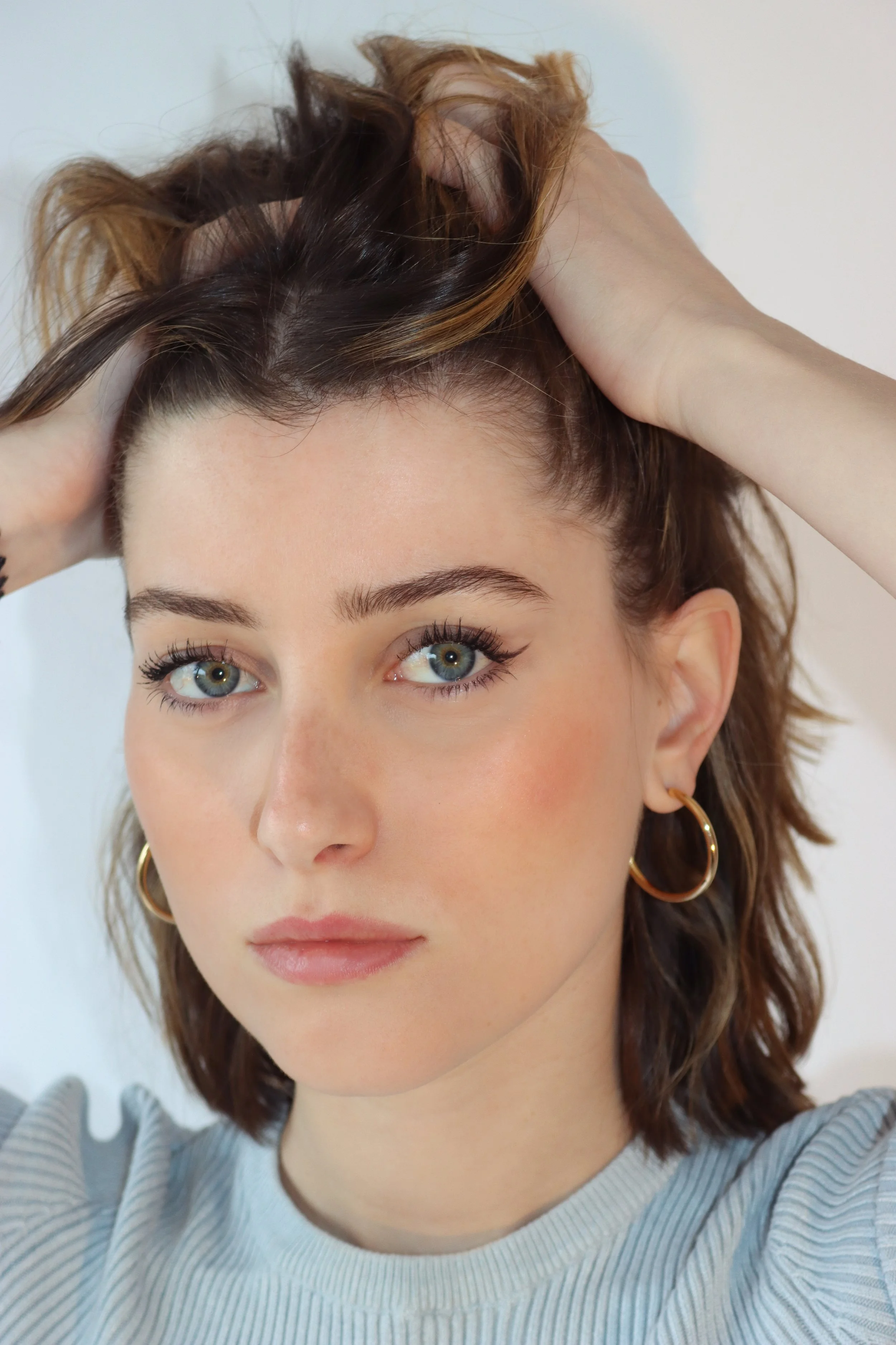 Jeune femme aux yeux verts, cheveux châtains avec des mèches blondes, portant de gros anneaux d'oreilles, pose avec une main dans ses cheveux, portant un pull gris à rayures, fond blanc.