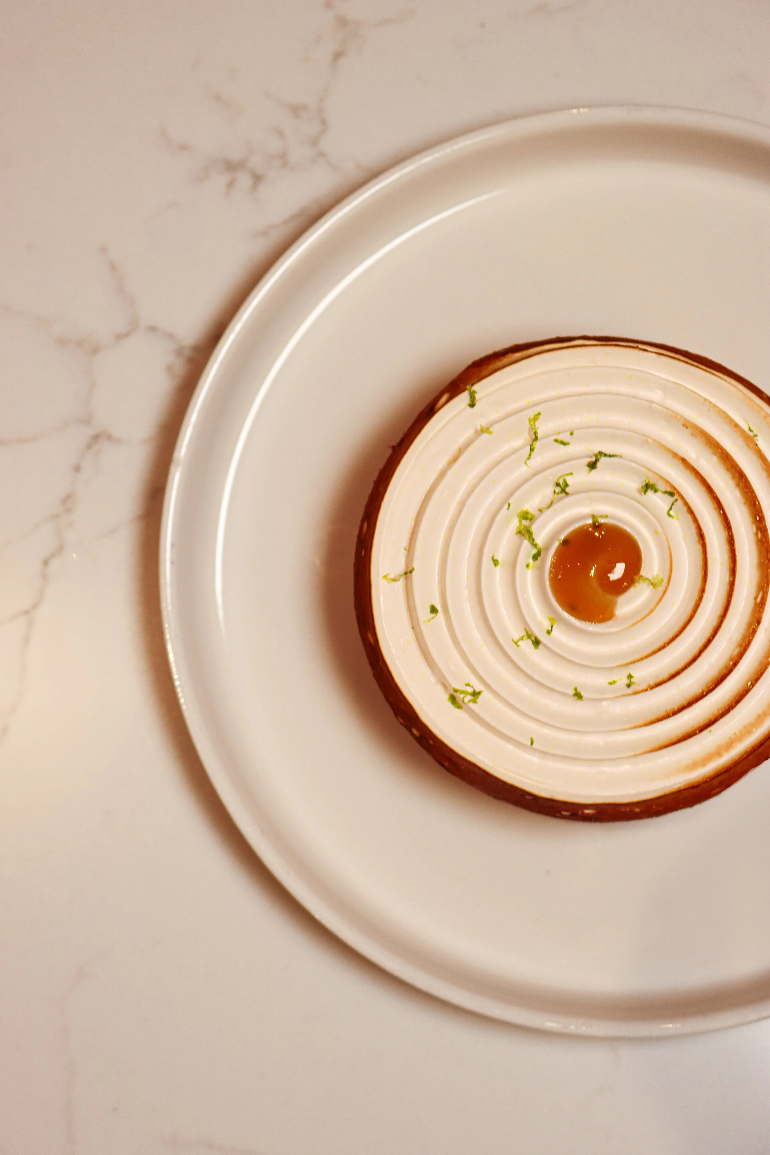 Gâteau avec un design géométrique en spirale, décoré de zestes de citron vert et d'un filet de caramel, présenté sur une assiette blanche.