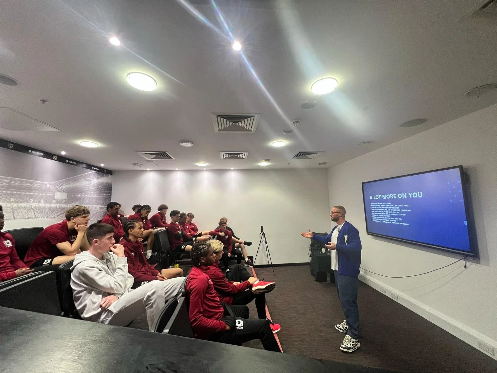 A group of young athletes in red and black sportswear listening to a man in a blue jacket giving a presentation in a conference room. The room has a large screen displaying a slide titled 'A LOT MORE ON YOU' and a wall mural of a sports stadium.