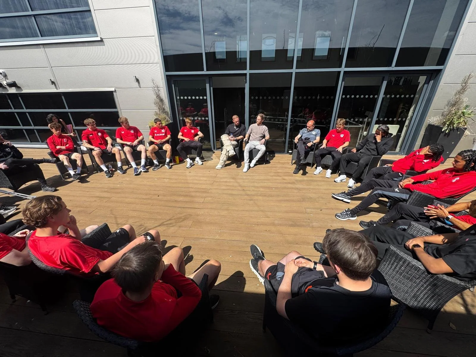 Group of athletes and staff sitting in a circle on a wooden deck outside a modern building, engaged in discussion.