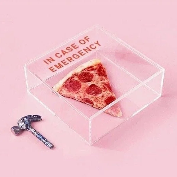 Une tranche de pizza au pepperoni dans une boîte en plastique claire avec l'inscription « IN CASE OF EMERGENCY » en rouge, placée sur une surface rose, à côté d'un marteau.