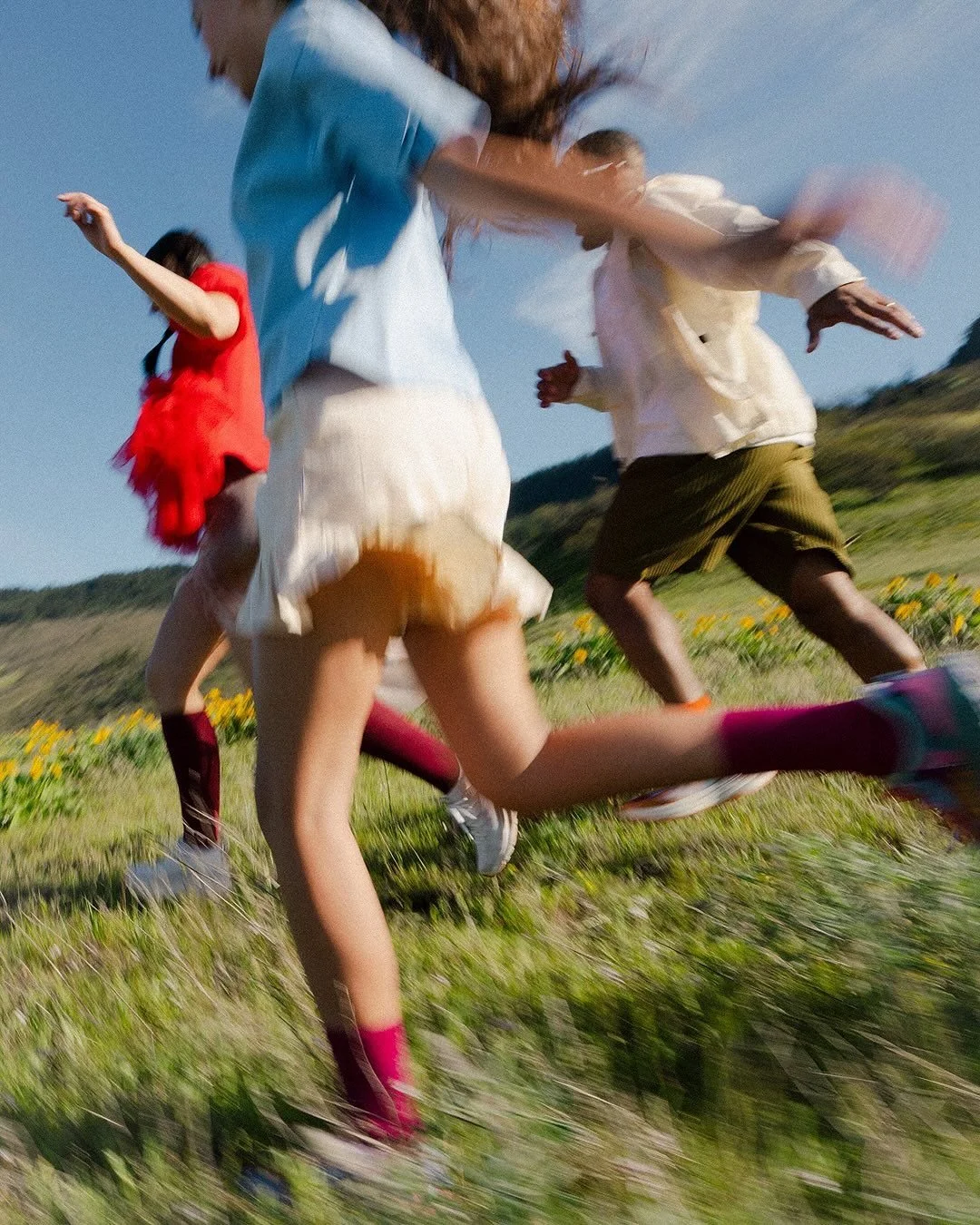 Trois enfants courant dans un champ avec des montagnes en arrière-plan, vêtus de vêtements colorés.