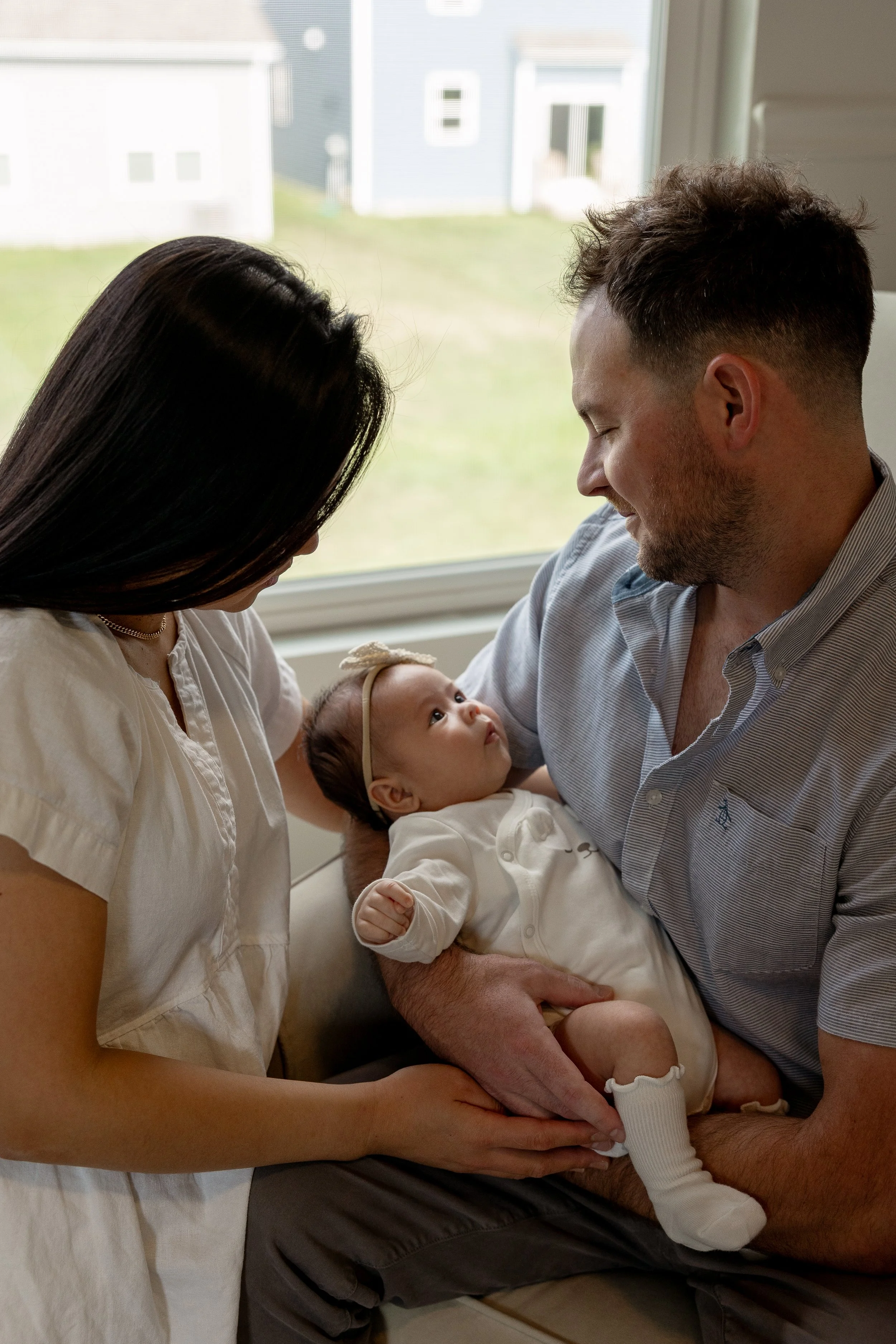 A family of three, including a mother, father, and a baby, sitting by a window indoors. The parents are looking at their baby, who is lying on the father's lap, dressed in cream-colored clothing and a headband. The mother is gently holding the baby's