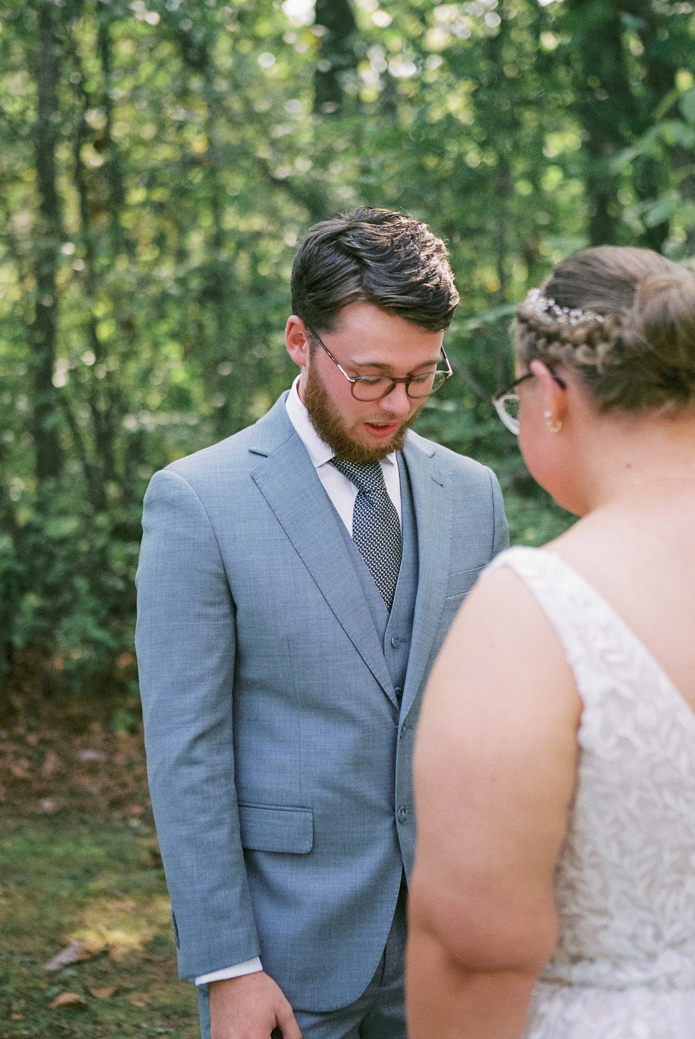 A man in a gray suit and glasses looks down with a solemn expression during an outdoor wedding ceremony. A woman in a lace wedding dress with glasses and a braided hairstyle is partially visible from the back.