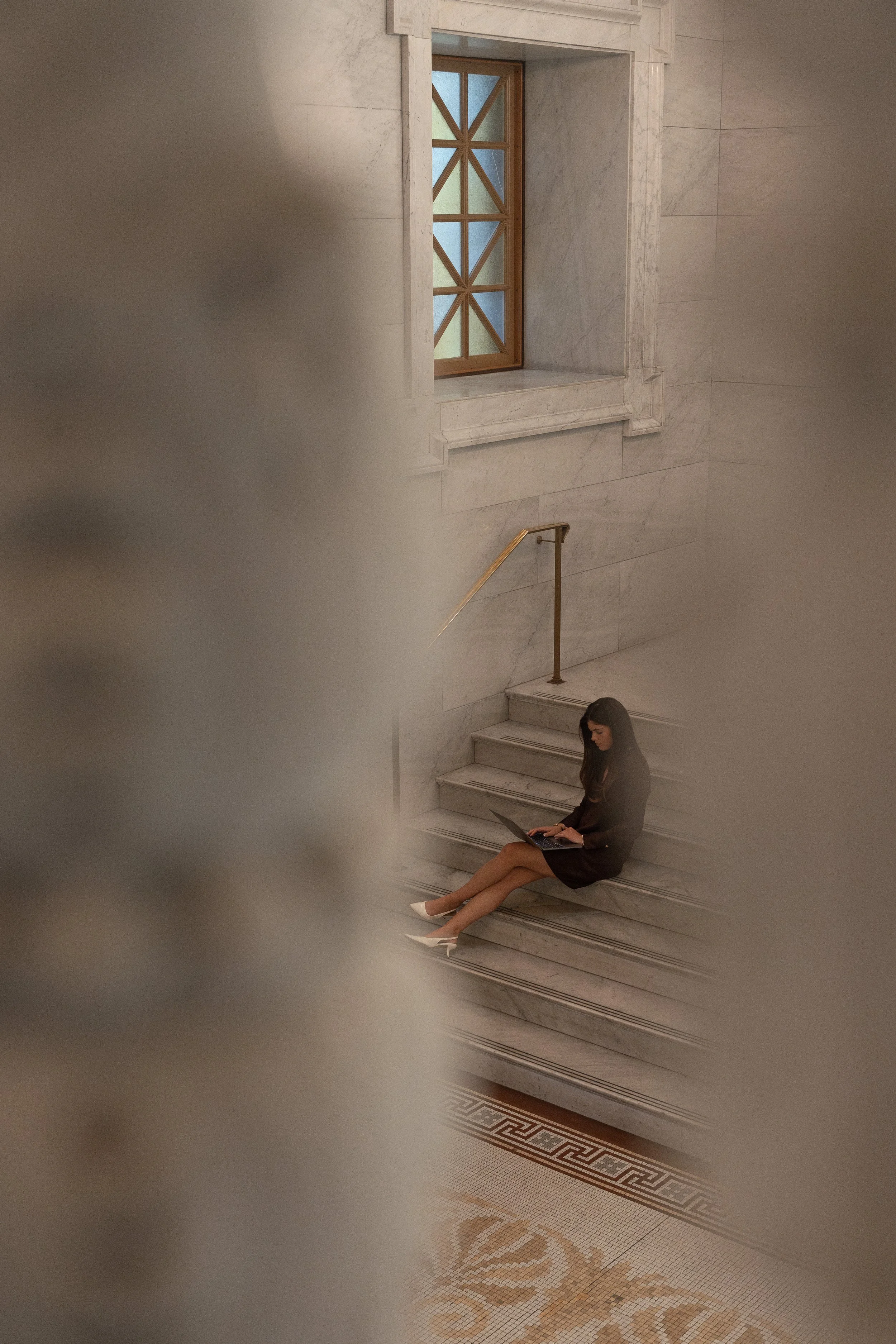 A woman in a black dress sitting on marble stairs inside a building, working on her laptop, with a stained glass window in the background.