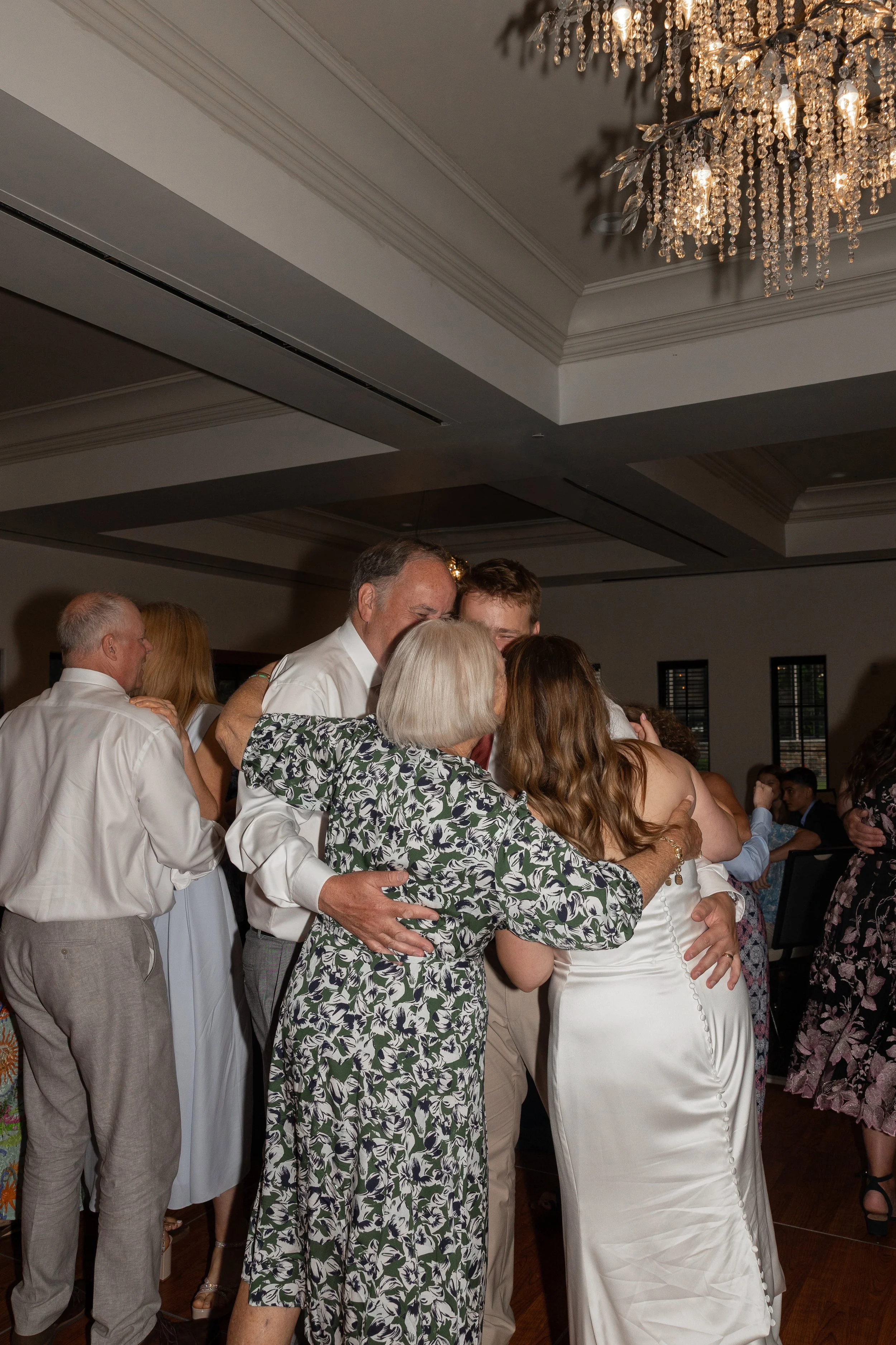 Group of people hugging at a wedding reception in a decorated indoor venue with a chandelier overhead.