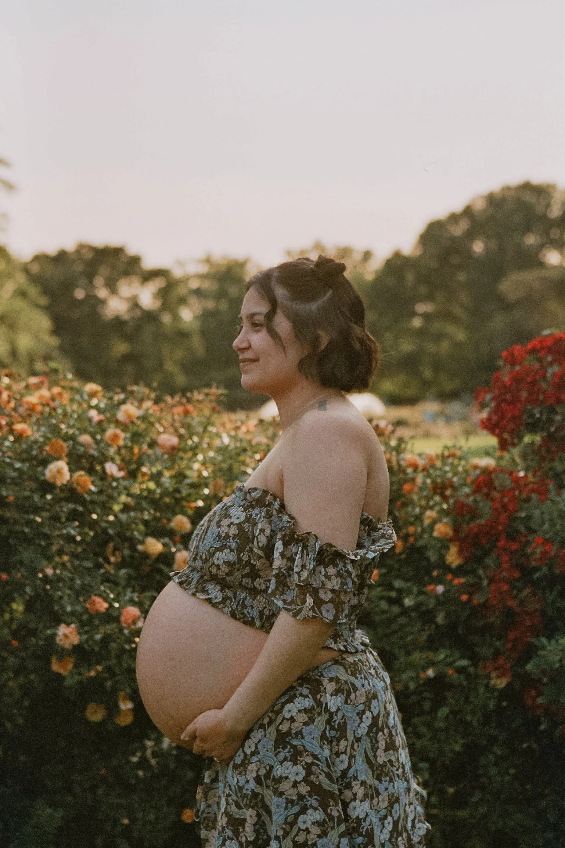 Pregnant woman in floral dress standing in a garden with blooming flowers, smiling softly.