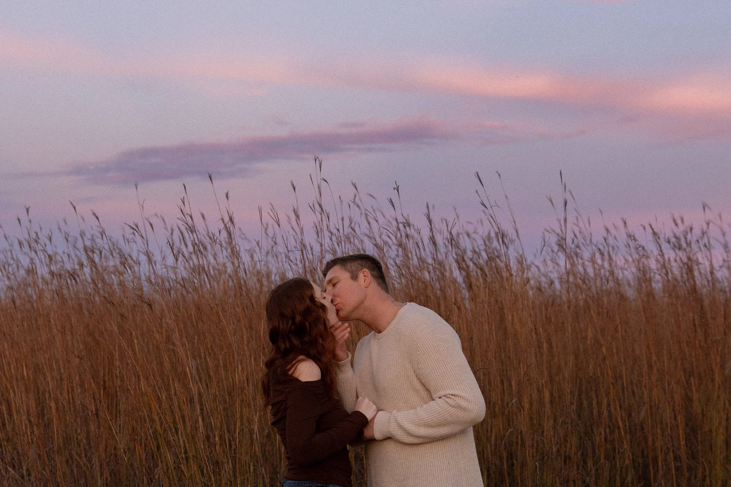 Couple kissing in a field of tall grass at sunset with pink and purple sky.