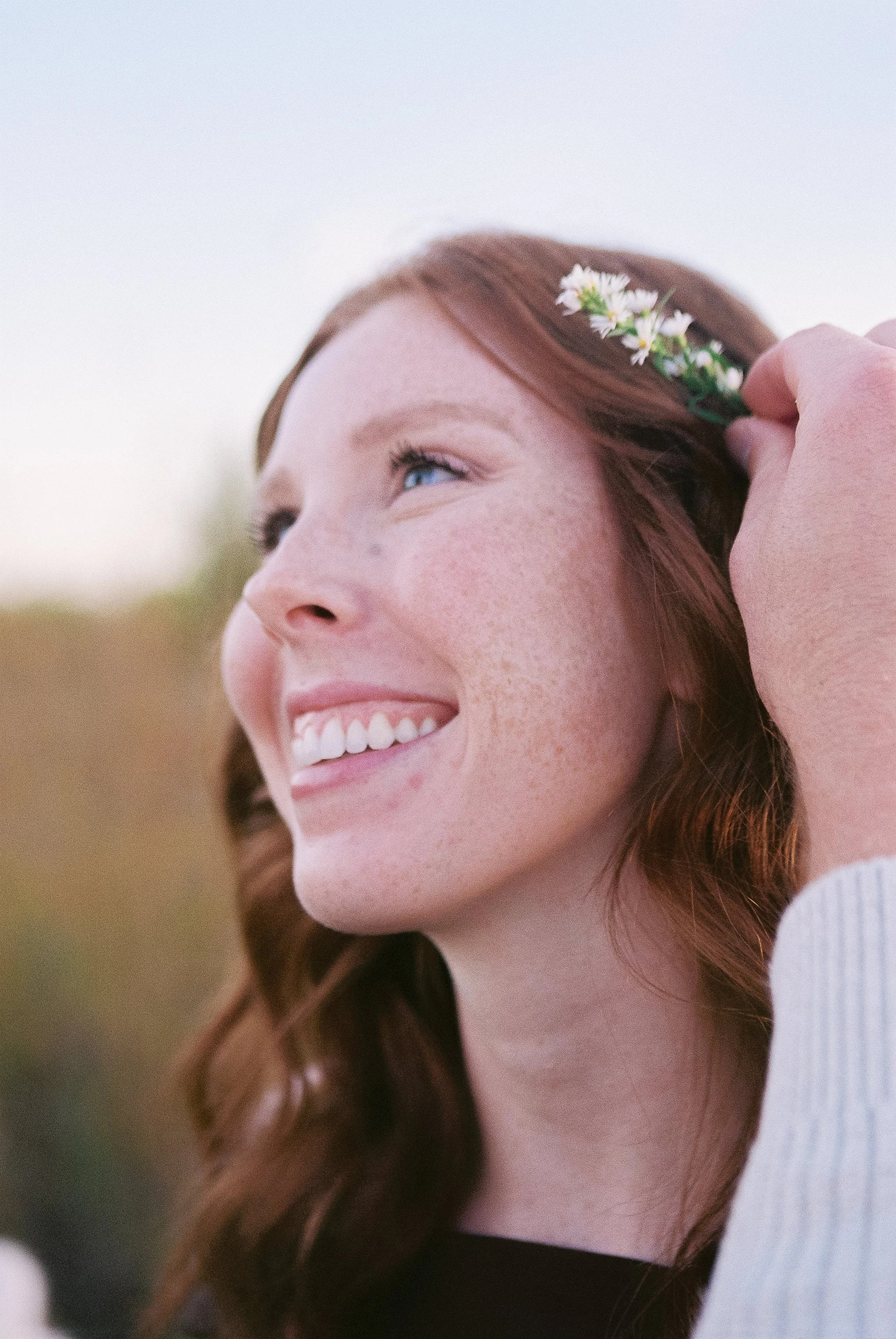 A young woman with red hair and freckles smiling while holding a small bouquet of white flowers near her ear outdoors during daylight.