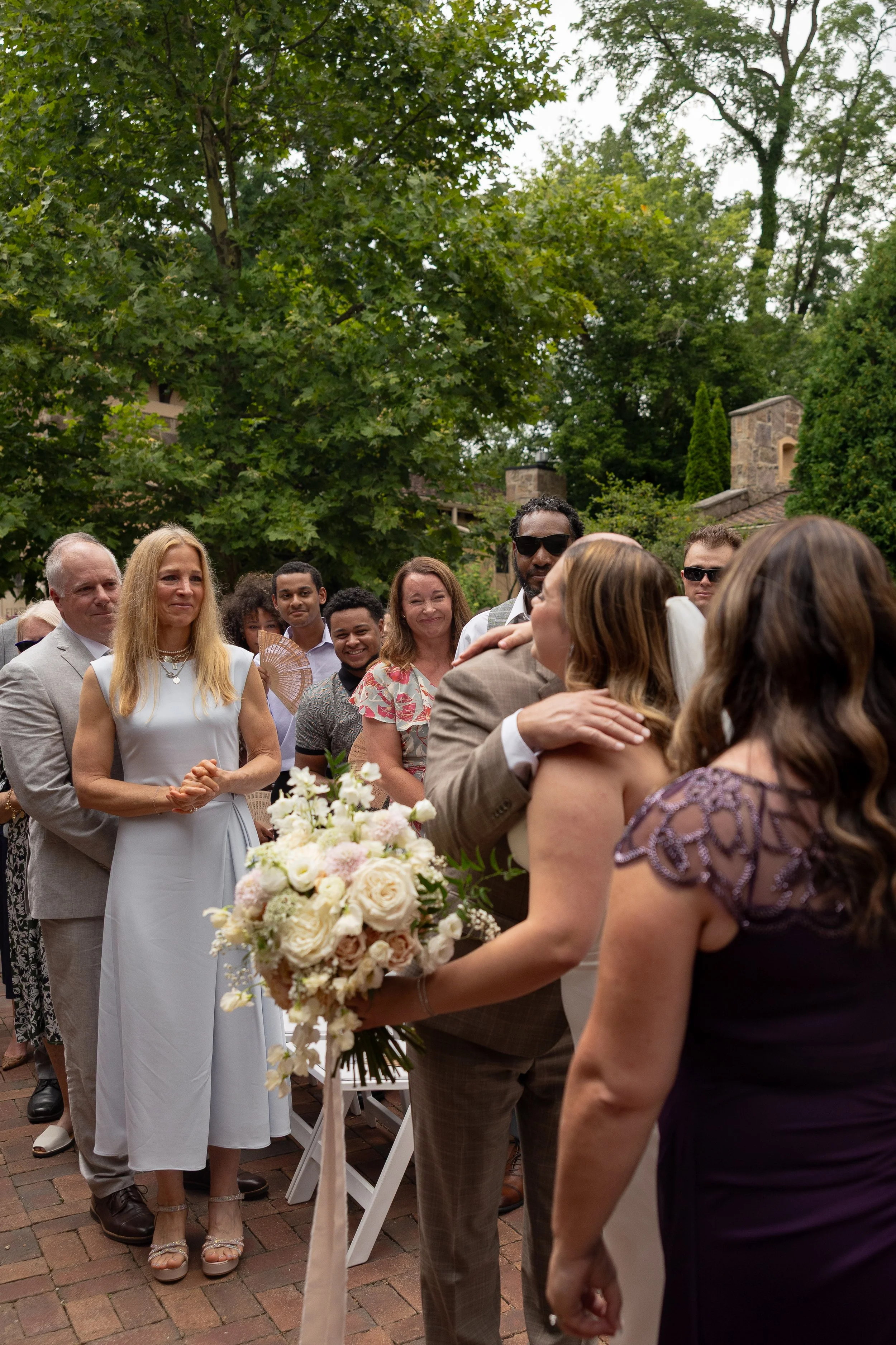 People at a wedding outdoors, embracing, with a bride holding a bouquet of flowers, others watching, under trees and a cloudy sky.