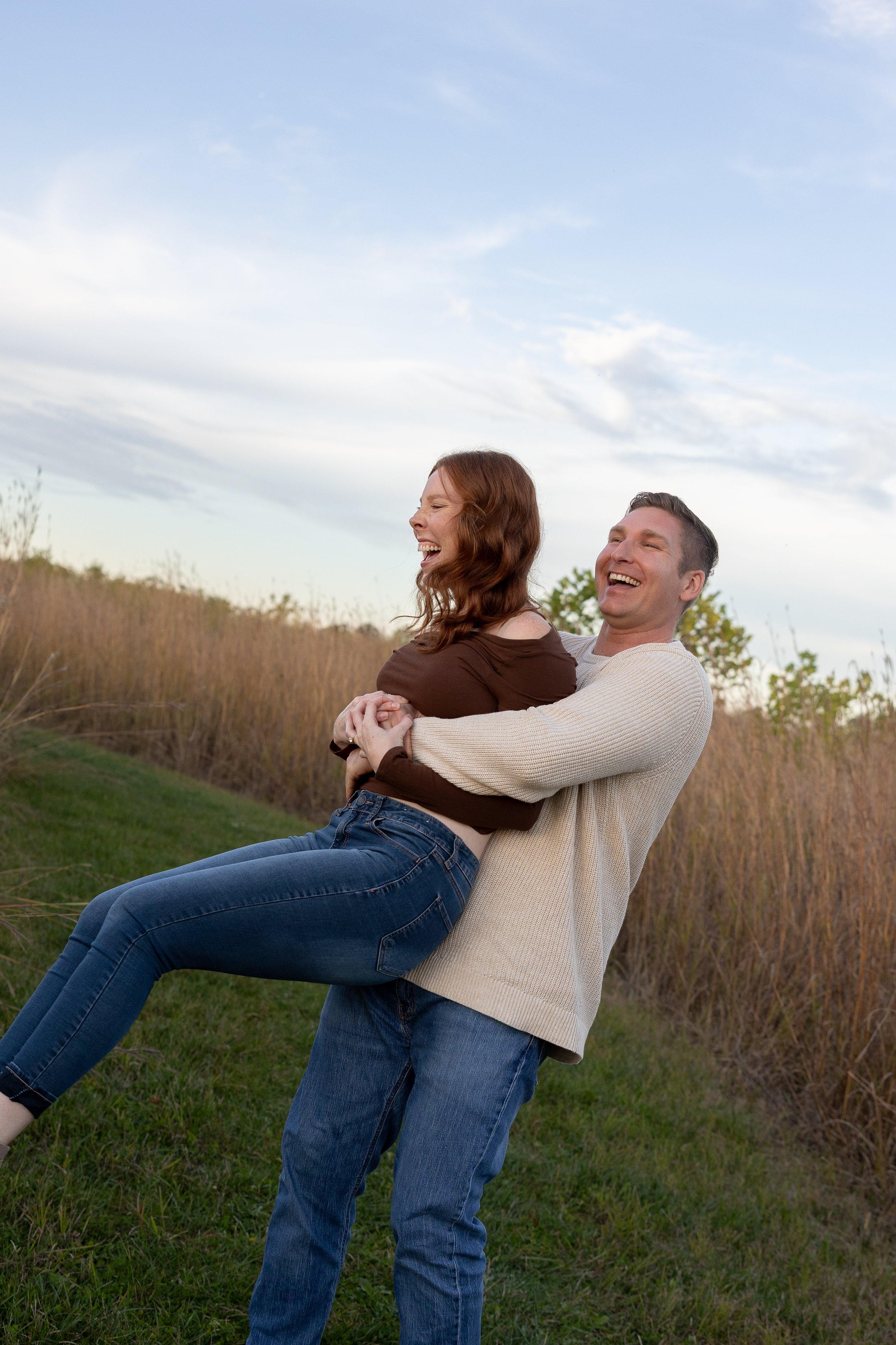 A couple is outdoors, with the man holding the woman in his arms, both laughing and enjoying each other's company.