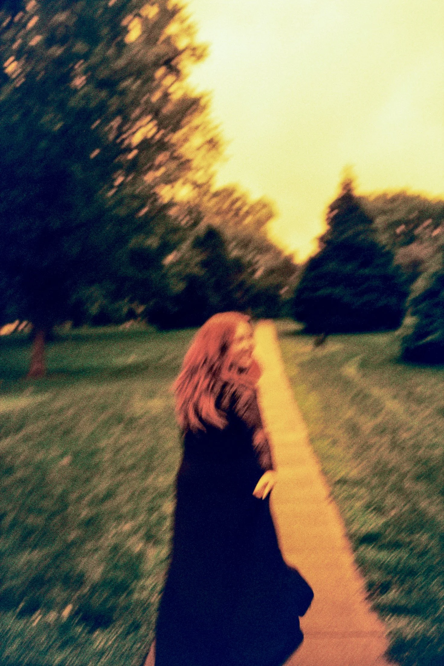 A woman with long hair walking along a park sidewalk during sunset, with trees lining both sides and the sky glowing yellow.