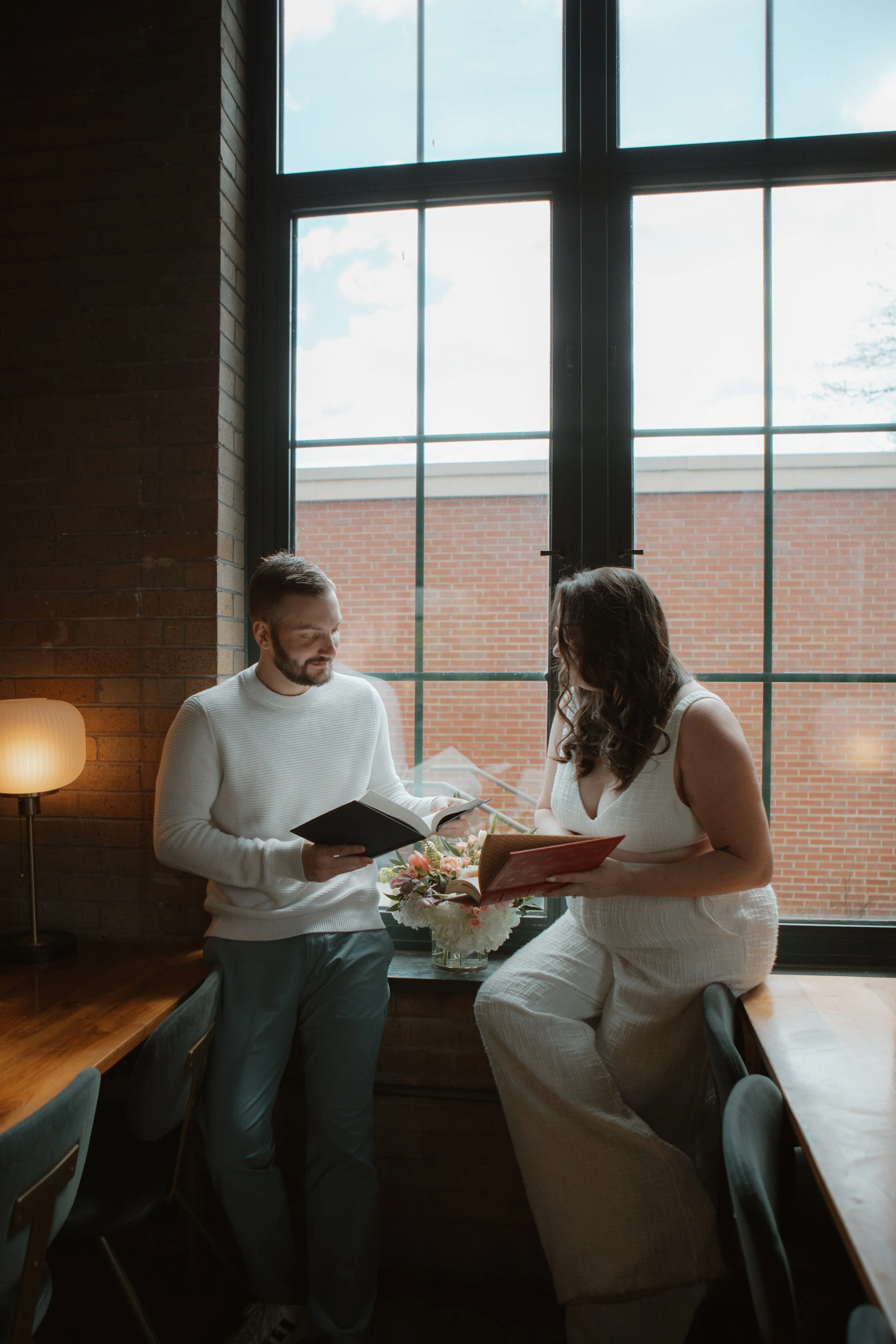 A man and woman are reading books and talking in a cozy indoor setting near a large window, with a flower arrangement on the window sill.