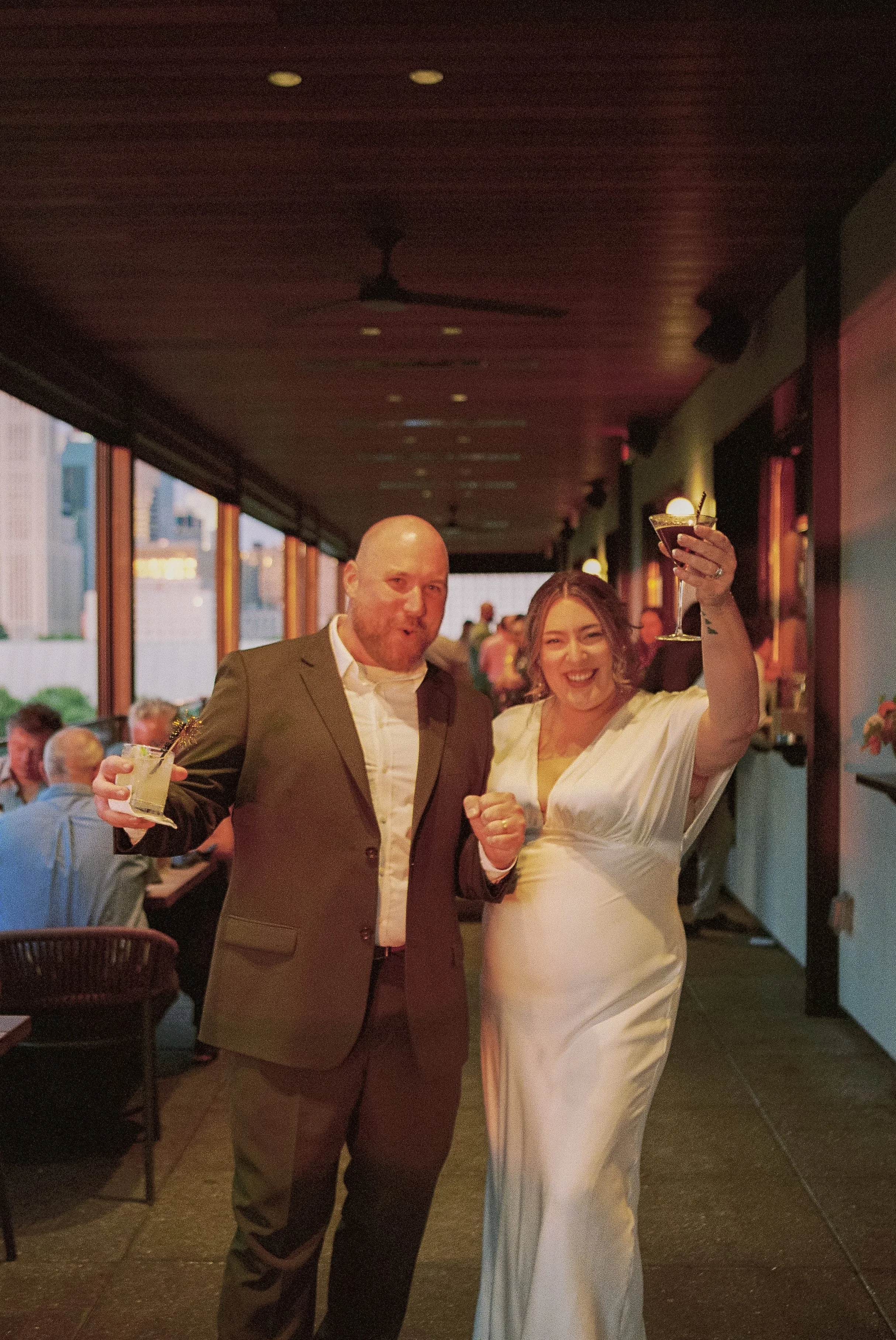 A man and woman celebrating at a social gathering, holding drinks and smiling in an indoor venue with large windows and city views.