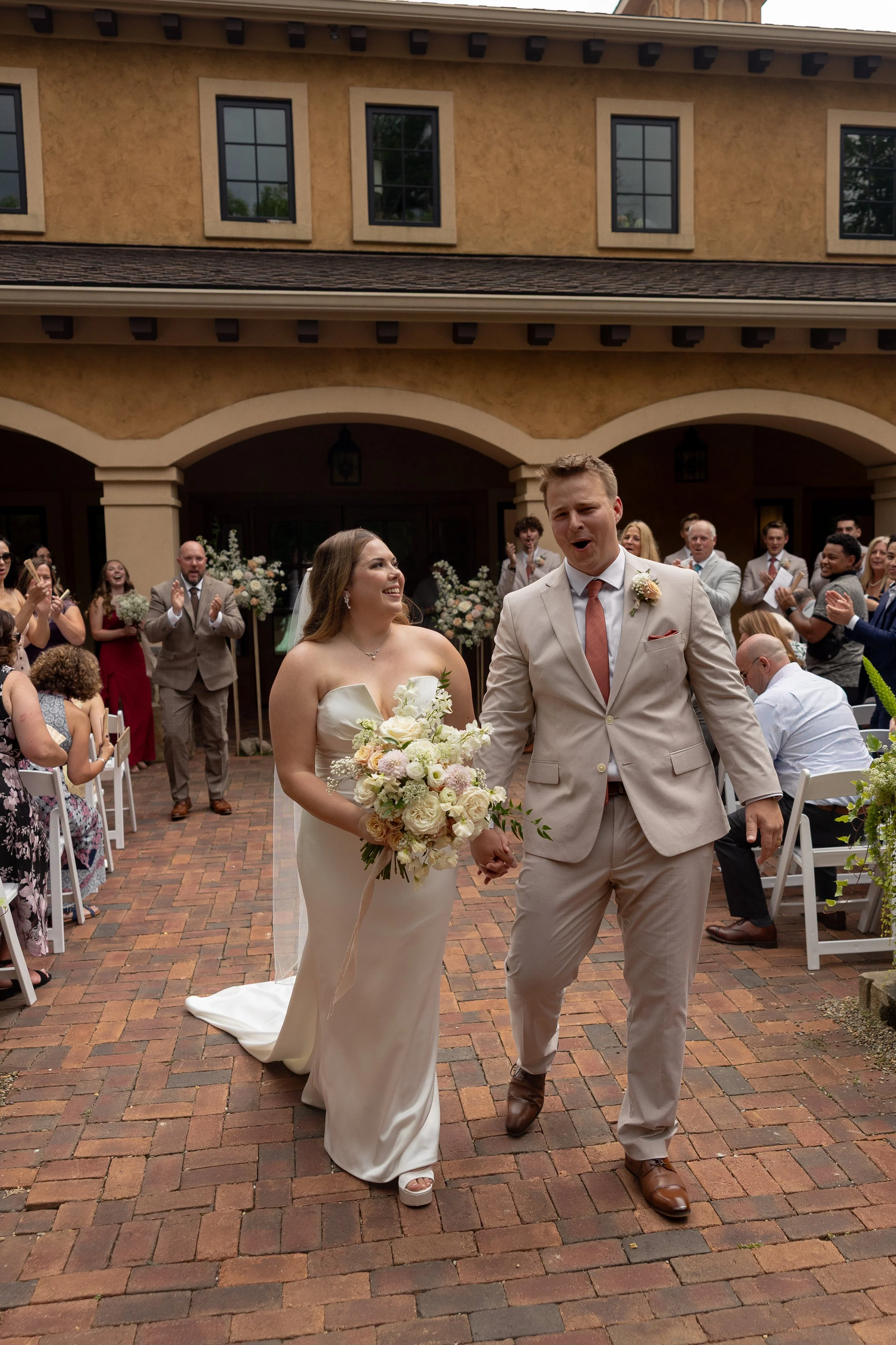 Bride and groom walking hand in hand at their outdoor wedding ceremony, with guests clapping and celebrating around them.