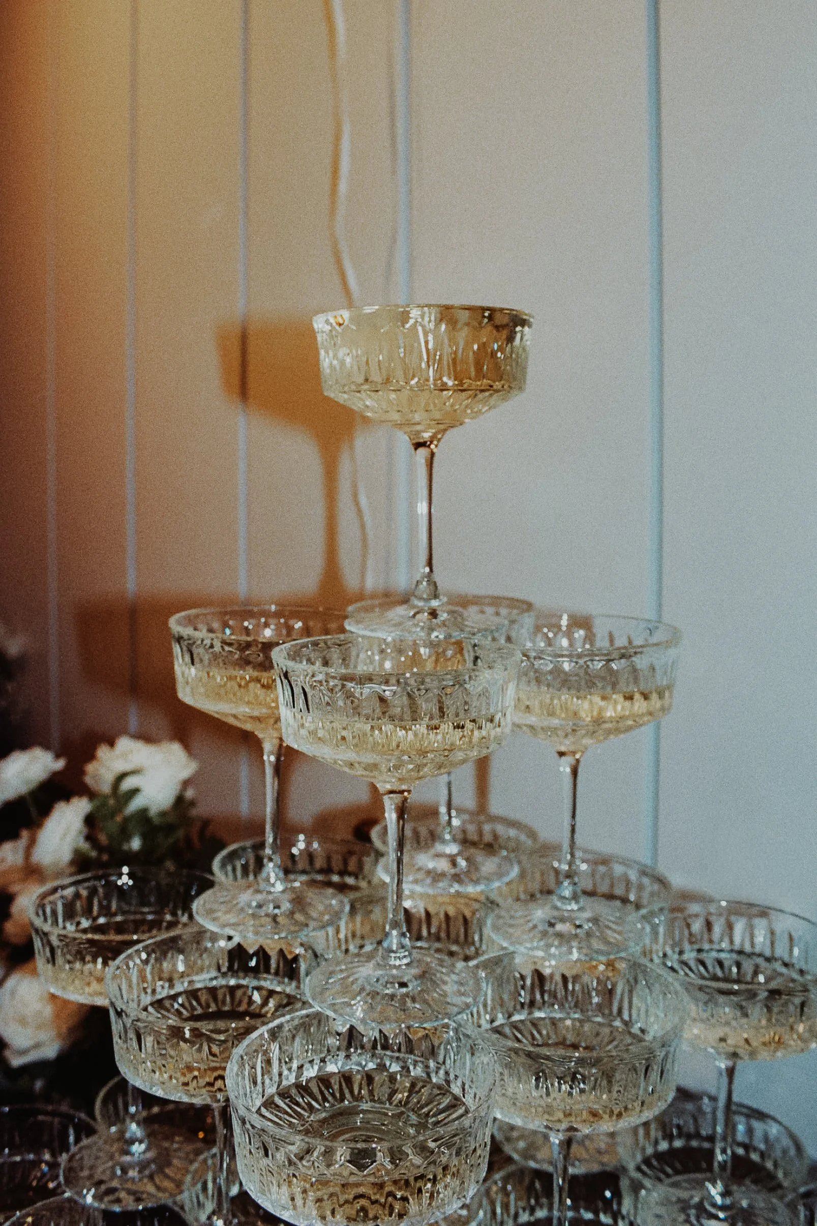 A pyramid of vintage glass champagne coupe glasses holding champagne, with some flowers and a textured wall in the background.