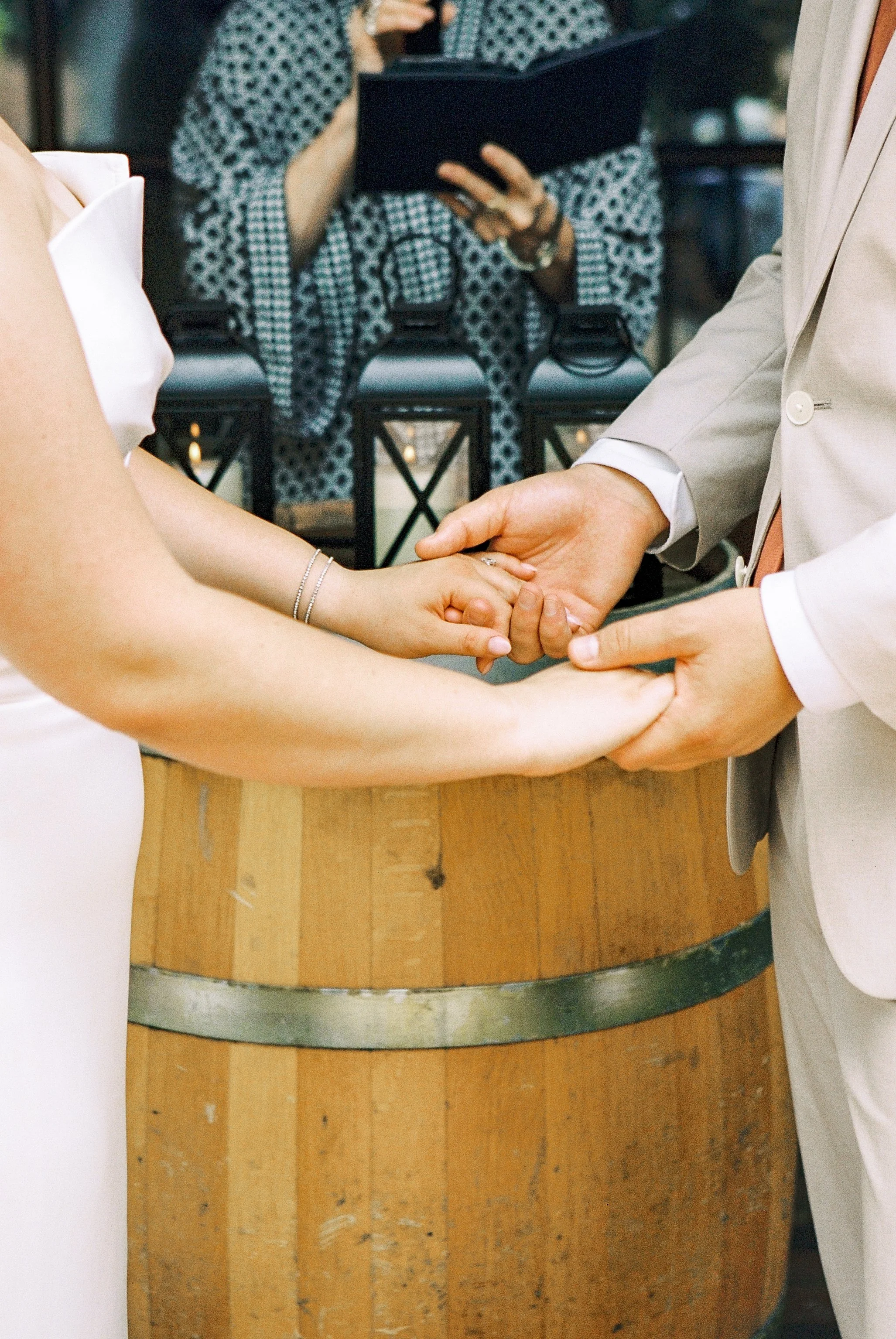 A couple holding hands during a wedding ceremony with a wooden barrel in the foreground.