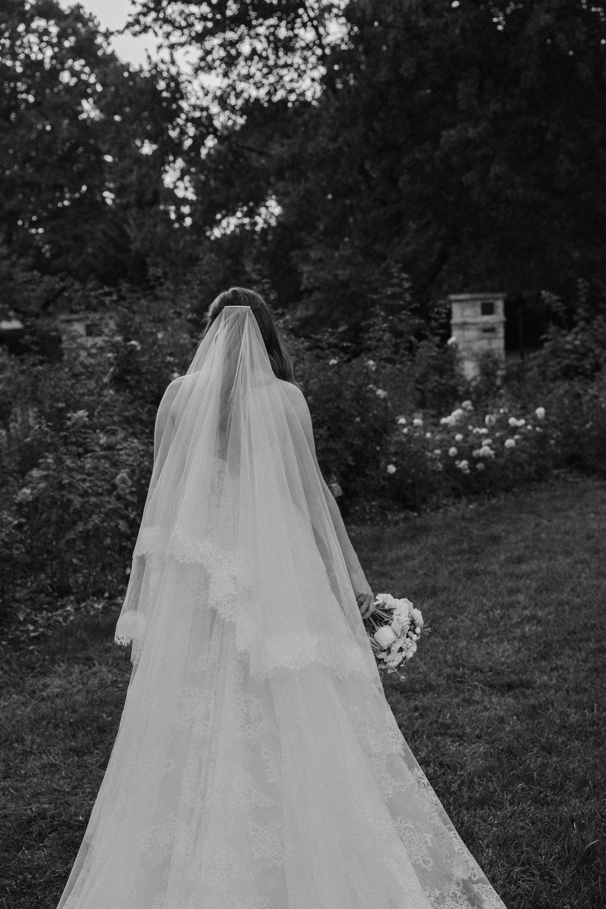 A woman in a wedding dress and veil holding a bouquet, walking away in a garden with trees and flowers, black and white photo.