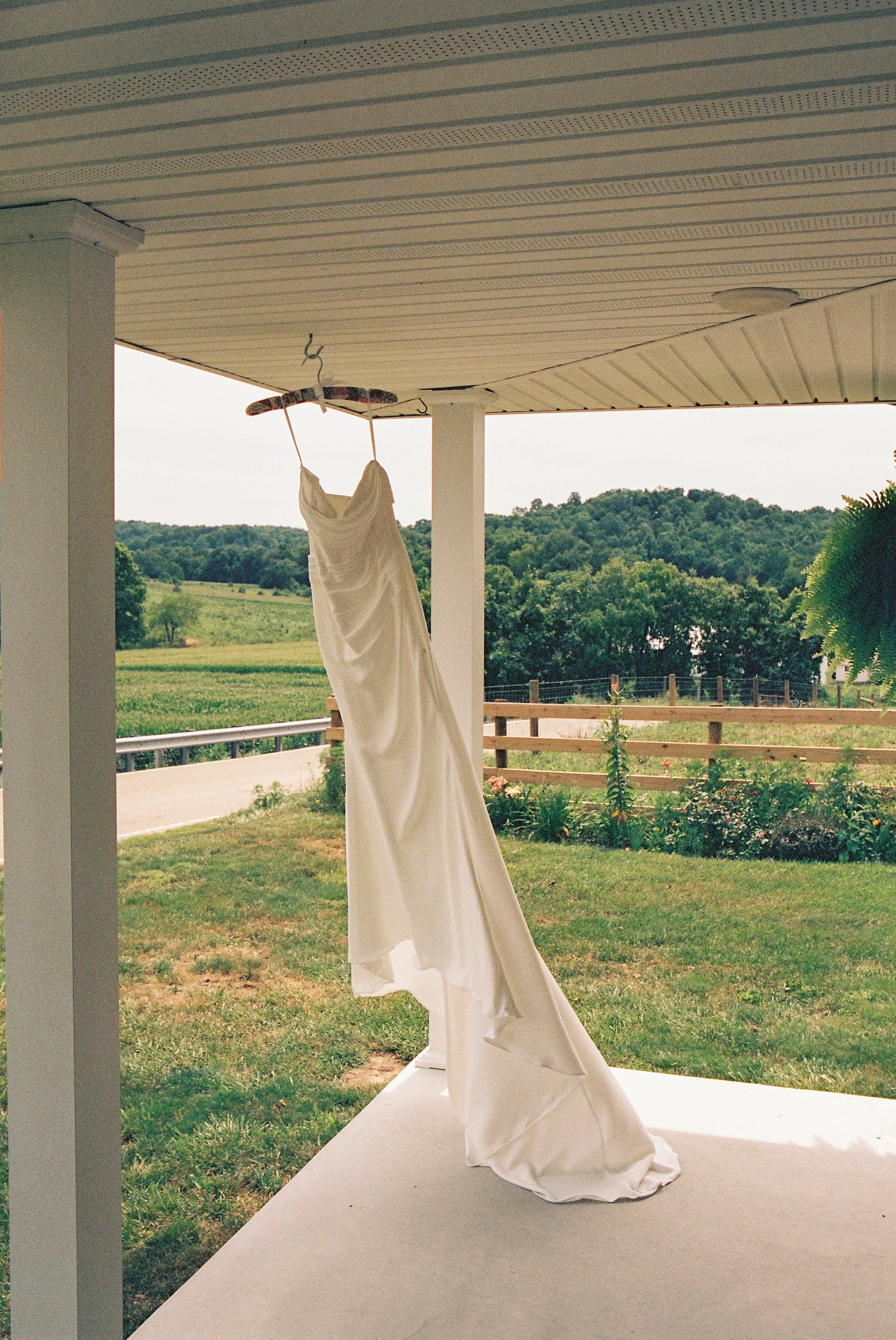 A white dress hanging on a hanger under a porch ceiling with a scenic view of green hills and trees in the background.