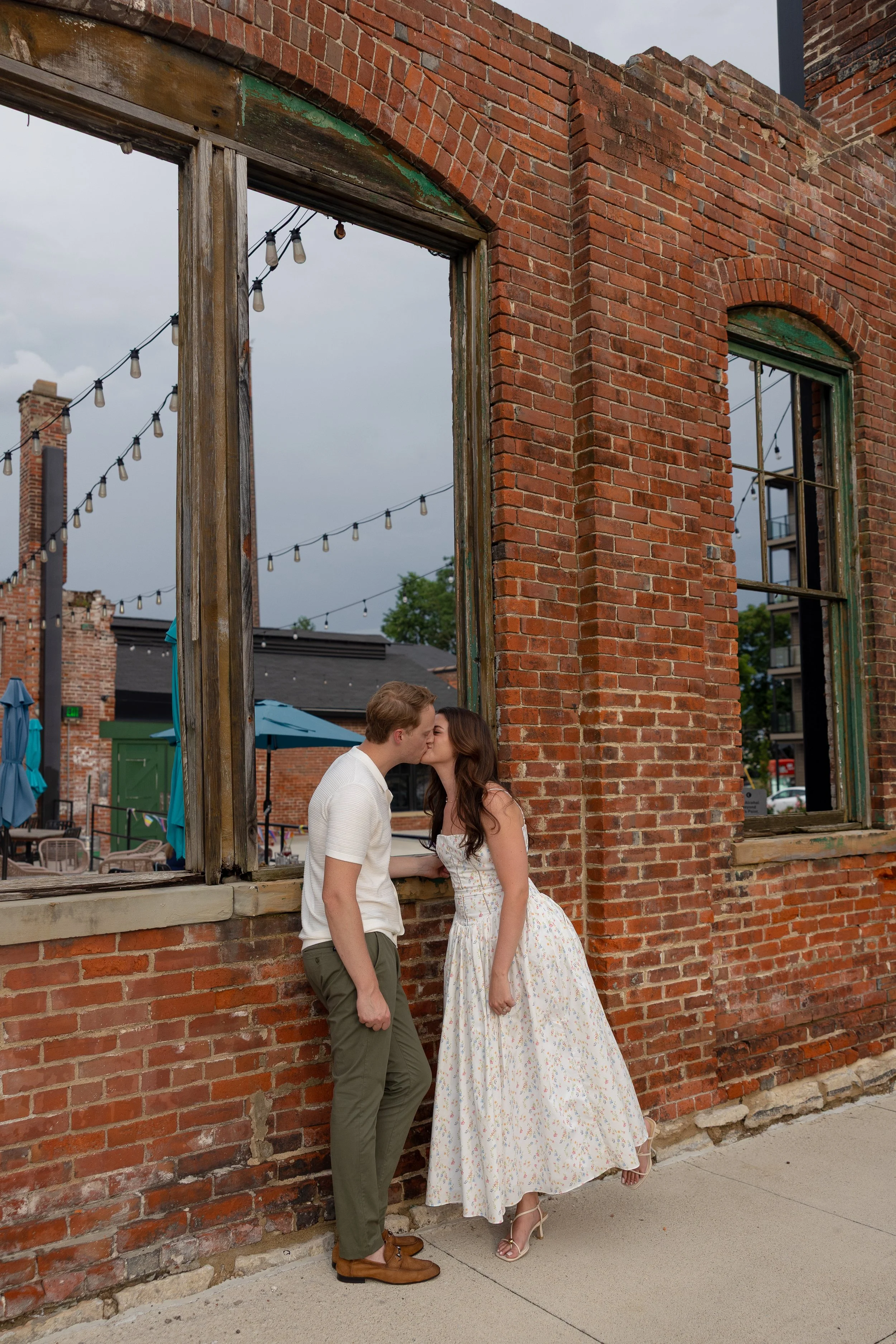 A couple kissing in front of an old brick building with large window frames and string lights hanging overhead.