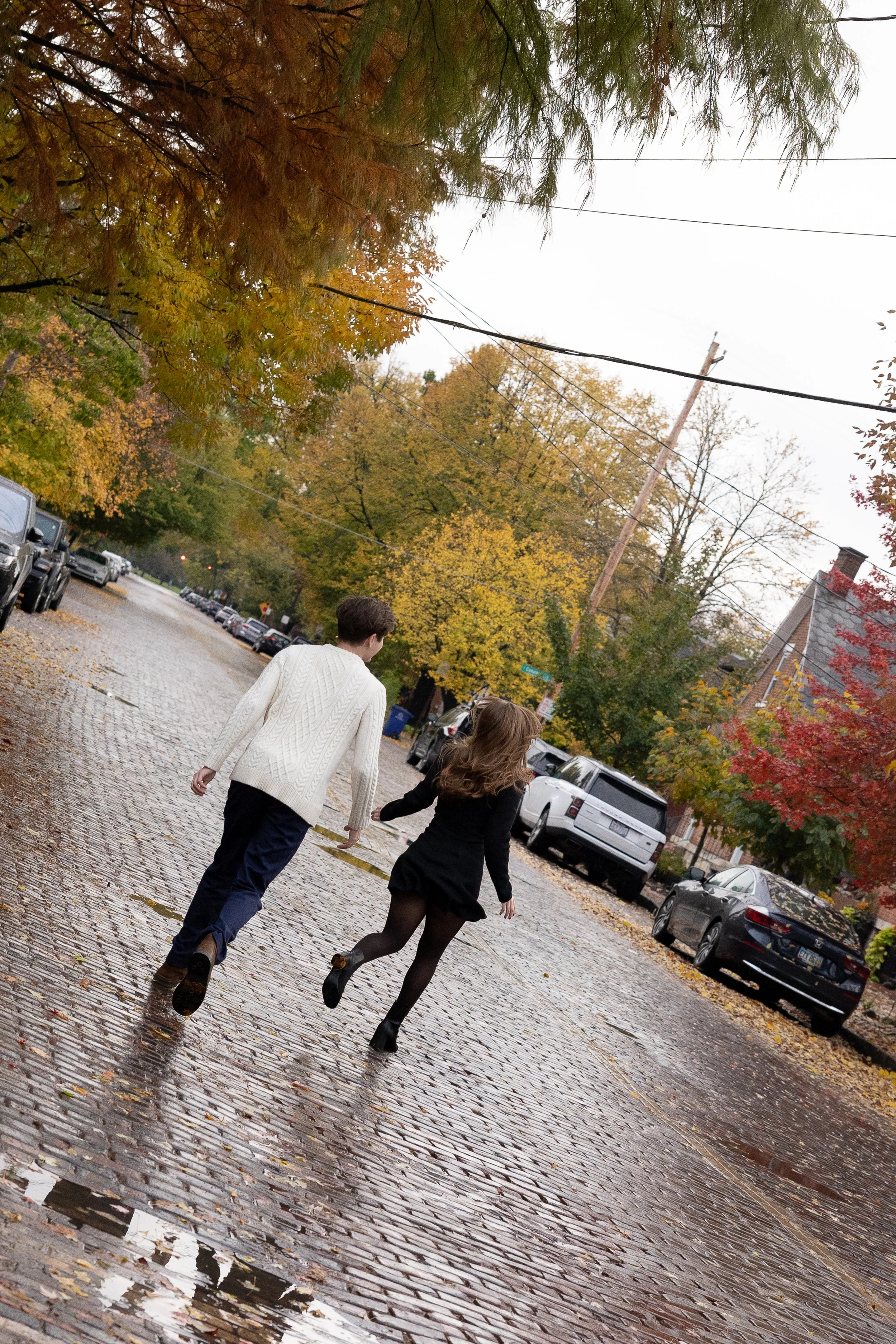 A young man and woman walking together on a wet cobblestone street during autumn, surrounded by colorful fall foliage and parked cars.