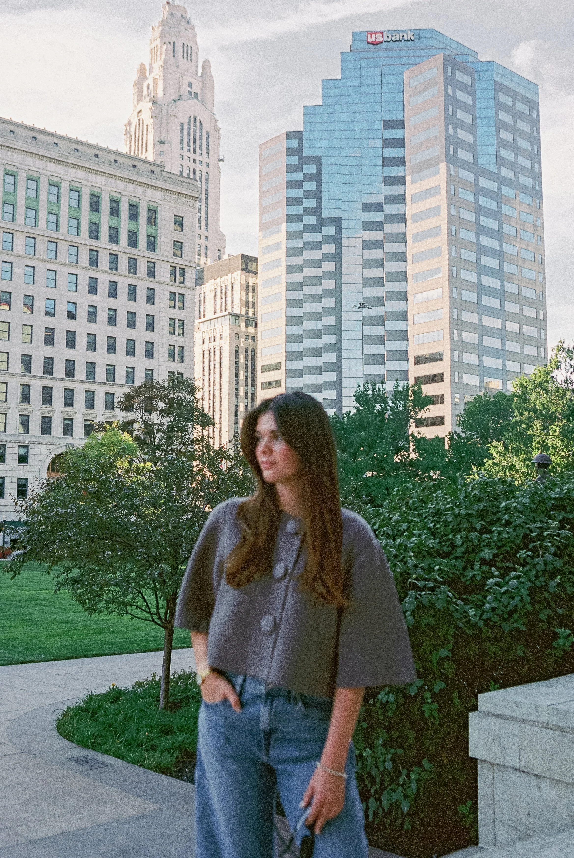 A young woman standing outdoors in an urban park with tall modern buildings in the background, including a US bank building.