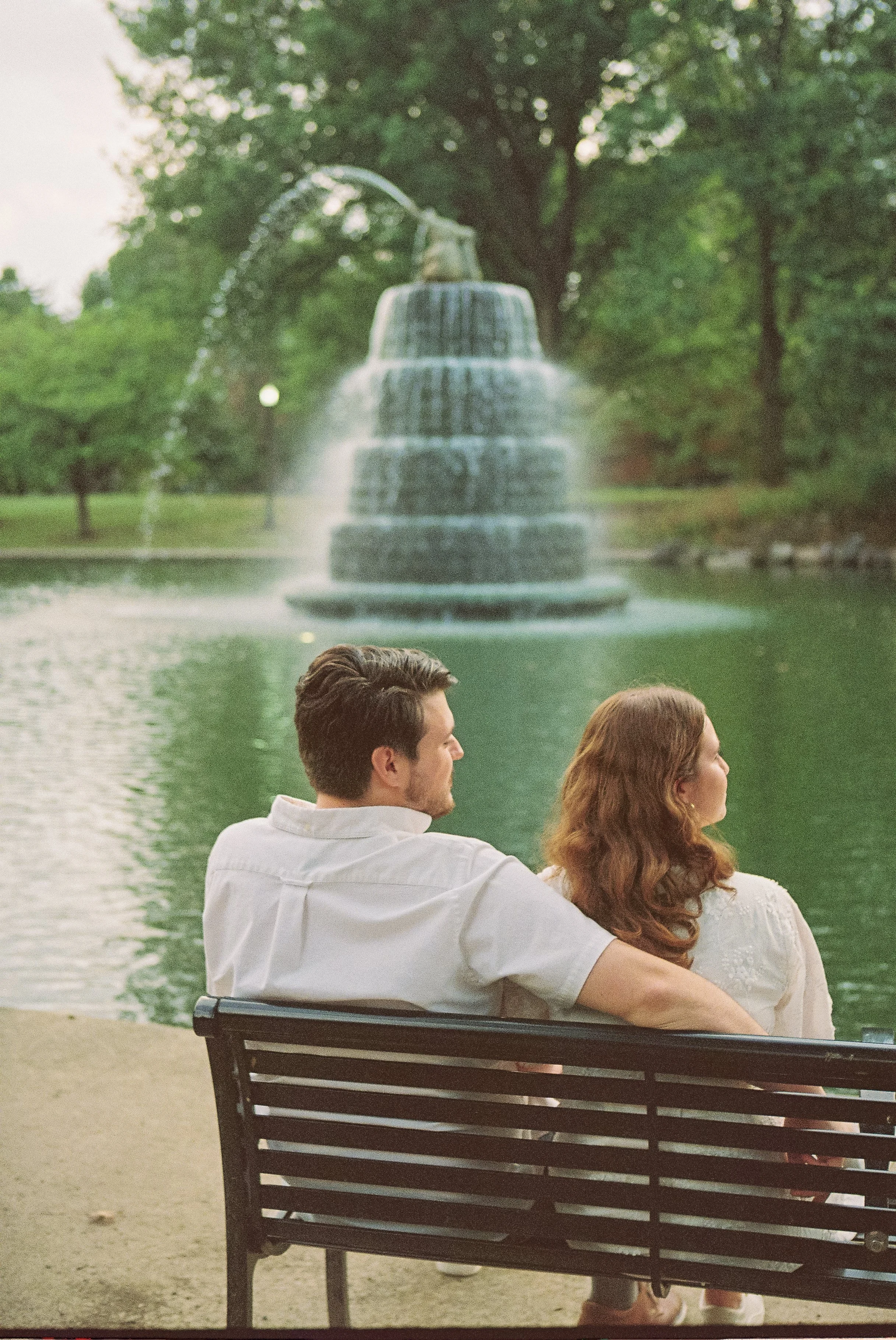 A man and woman sitting on a park bench by a pond with a fountain in the background, surrounded by trees.