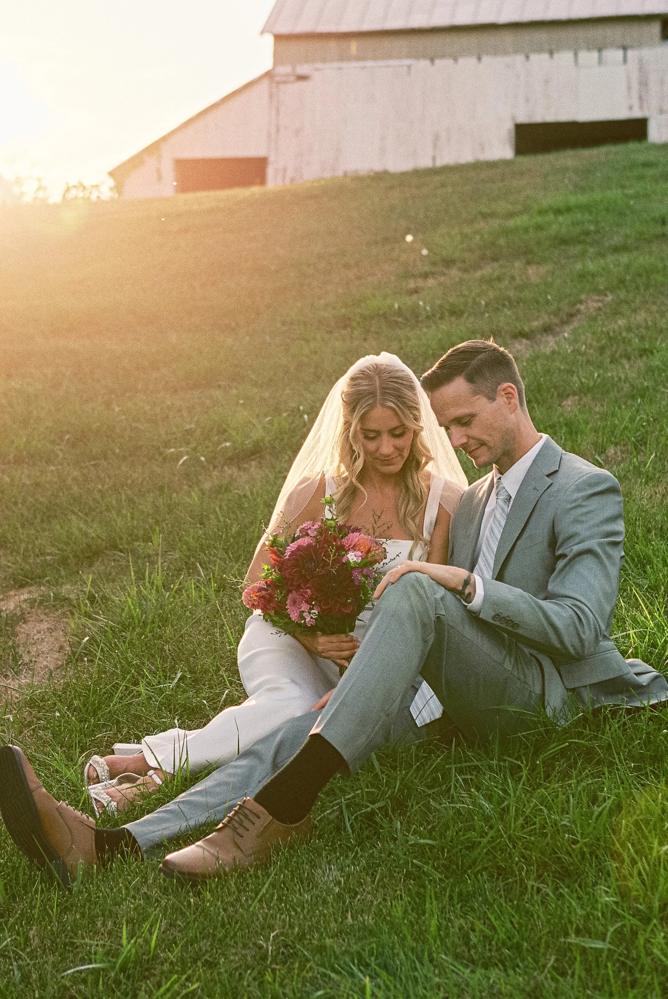 A bride and groom sitting on grass during sunset, with the bride holding a bouquet of pink and red flowers, both looking at each other's leg with smiles.