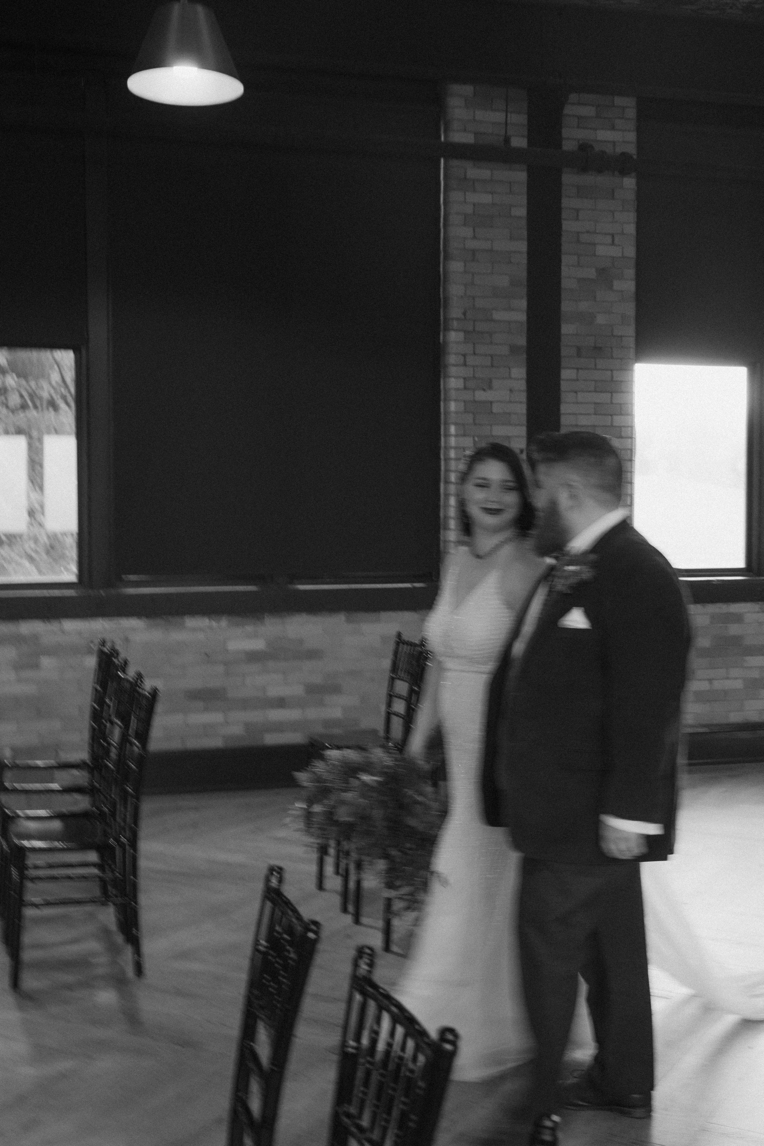 A bride and groom walking together at a wedding reception, with wedding attire and a floral bouquet, in a room with brick walls and large windows.