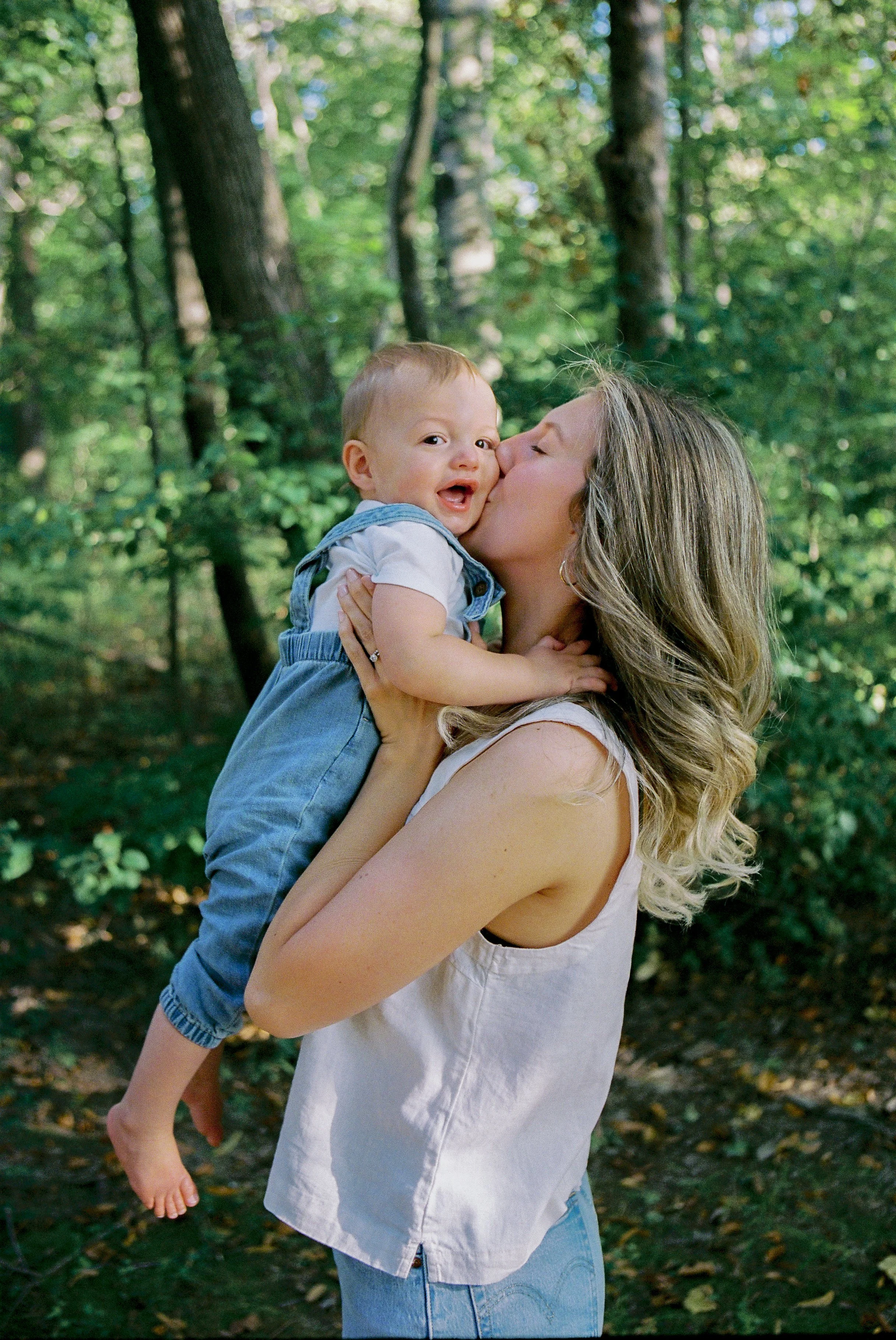 A woman with wavy blonde hair holds a young child outside in a wooded area, kissing him on the cheek as he smiles and looks at the camera.