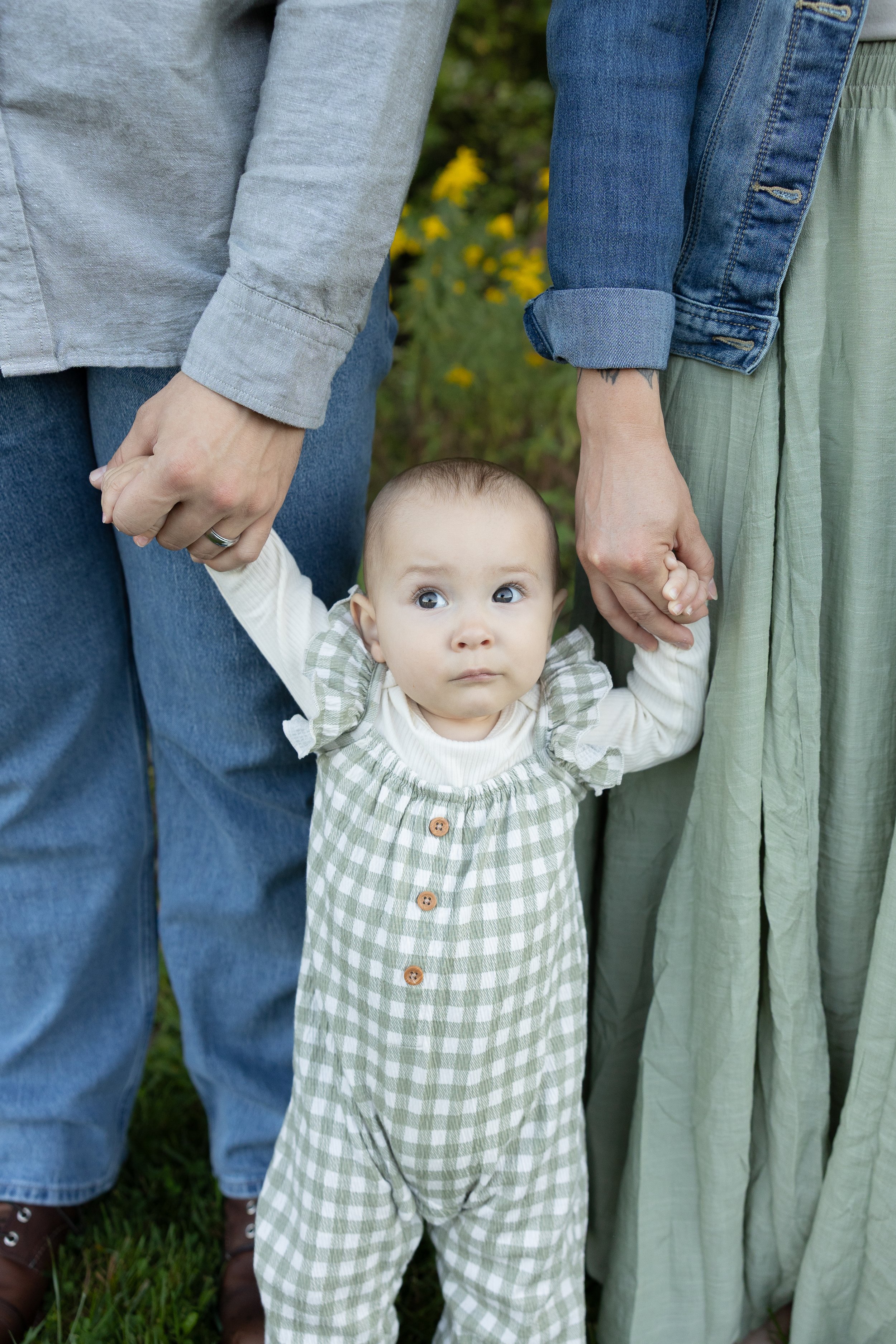 A baby holding hands with two adults outdoors, with yellow flowers and greenery in the background.