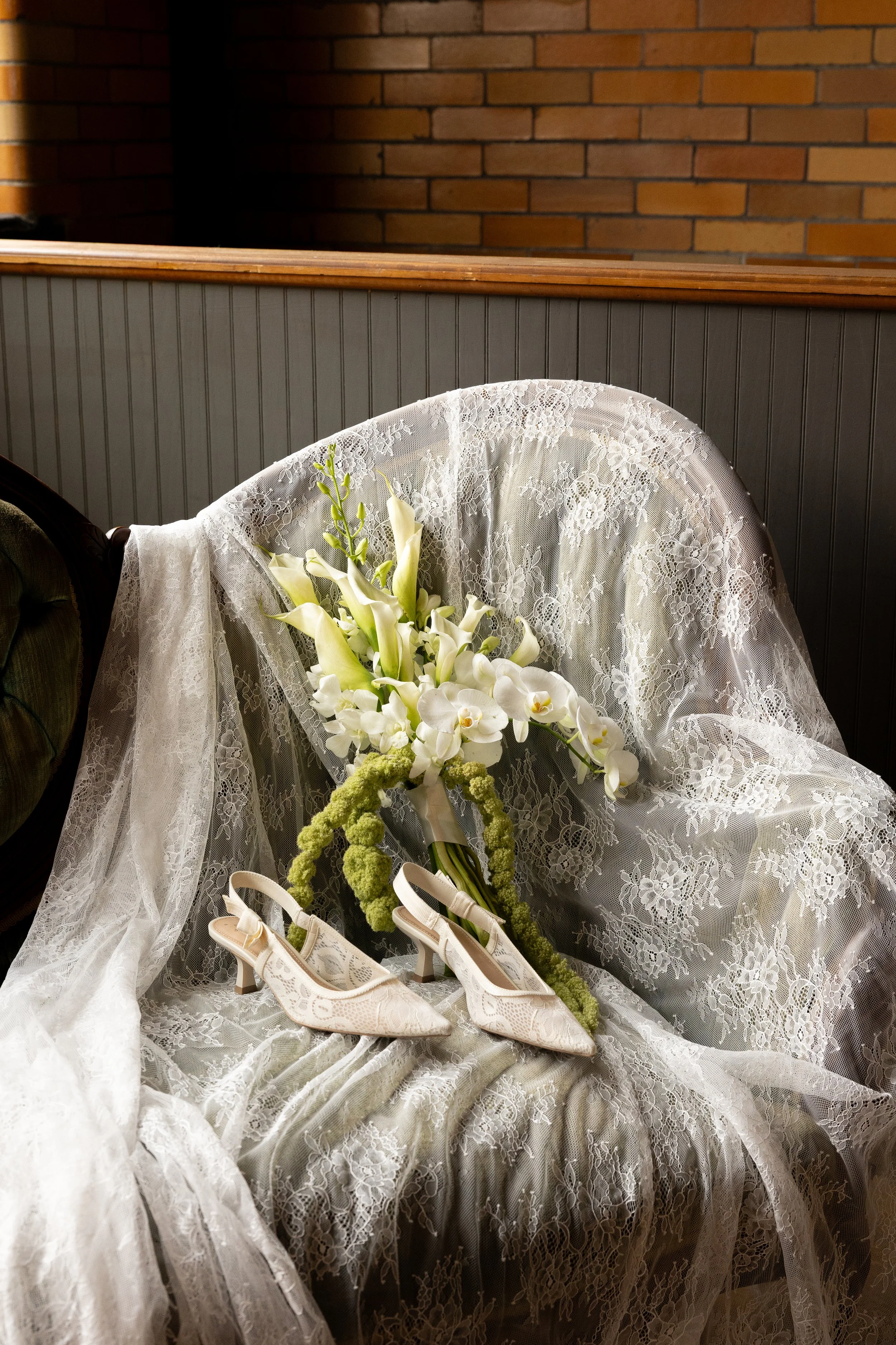 A bridal bouquet with white flowers and green accents, placed on a lace-covered chair with white lace shoes beside it.